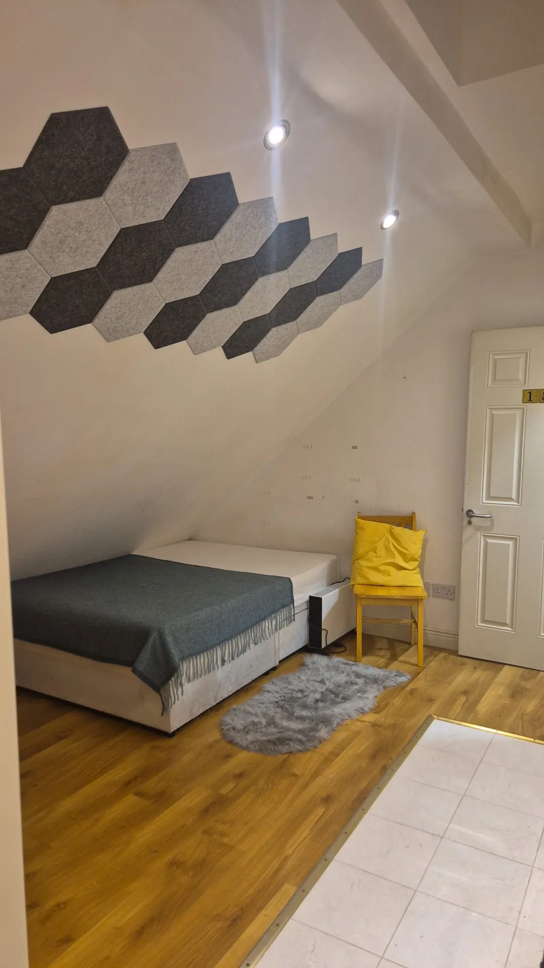 Bedroom with a single bed against a sloped ceiling, hexagonal acoustic panels on the ceiling, recessed spotlights, a yellow chair with cushion beside a door, and a small rug on wooden floor.