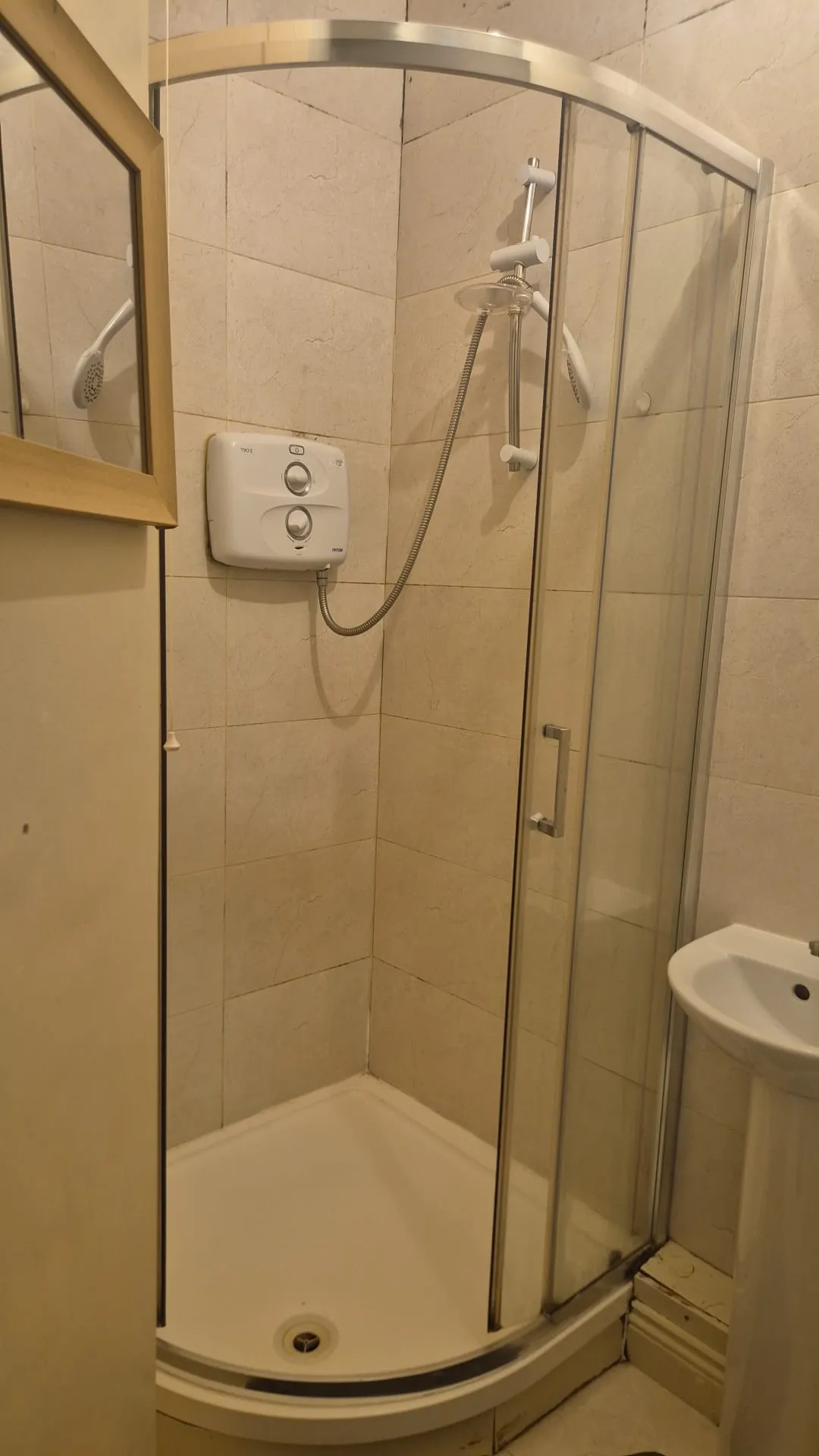 Small bathroom corner shower with curved glass doors, wall-mounted electric shower unit and handheld showerhead, tiled walls and a partial view of a pedestal sink and mirror.