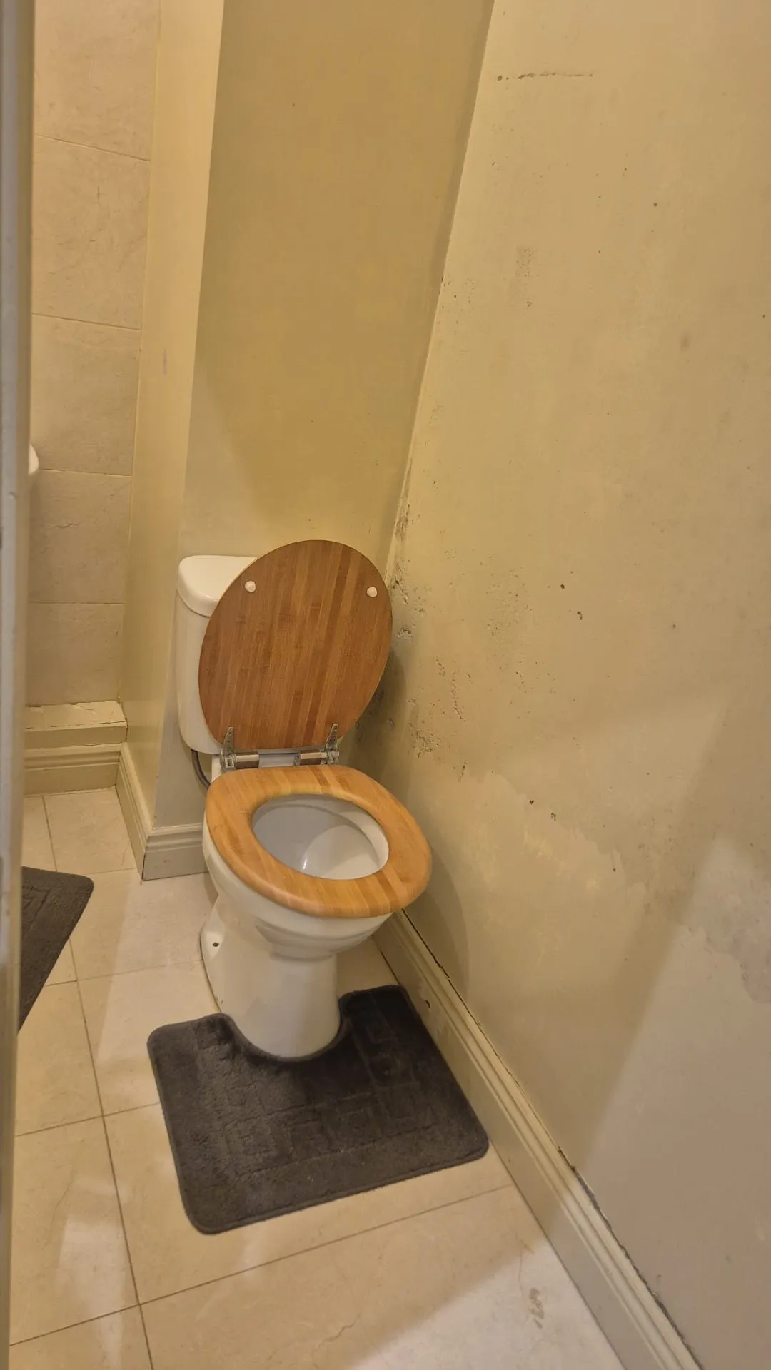 Small bathroom with a white toilet featuring a wooden seat, a dark floor mat, tiled floor, and a stained wall to the right.