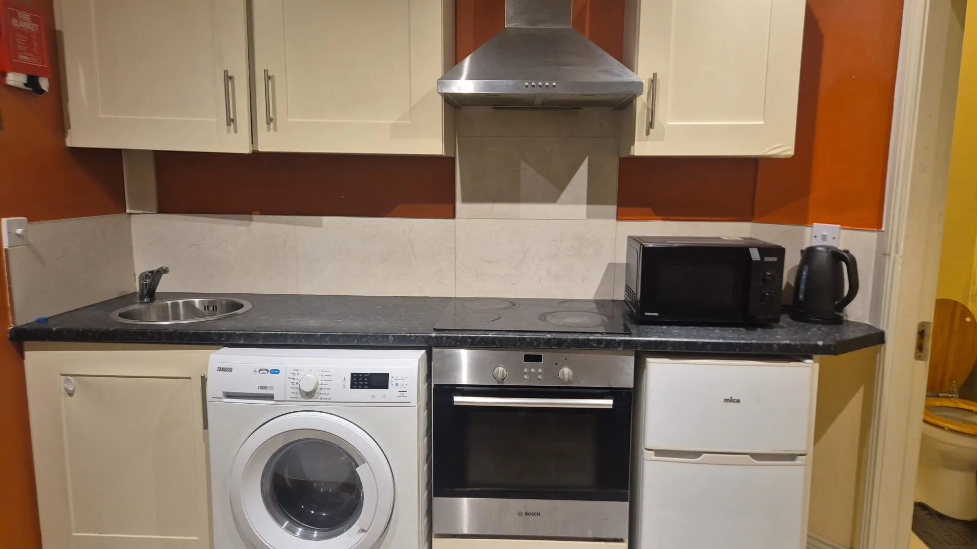 Kitchen countertop with cabinets, extractor hood, electric stovetop and oven, washing machine, microwave, kettle, sink and small fridge.