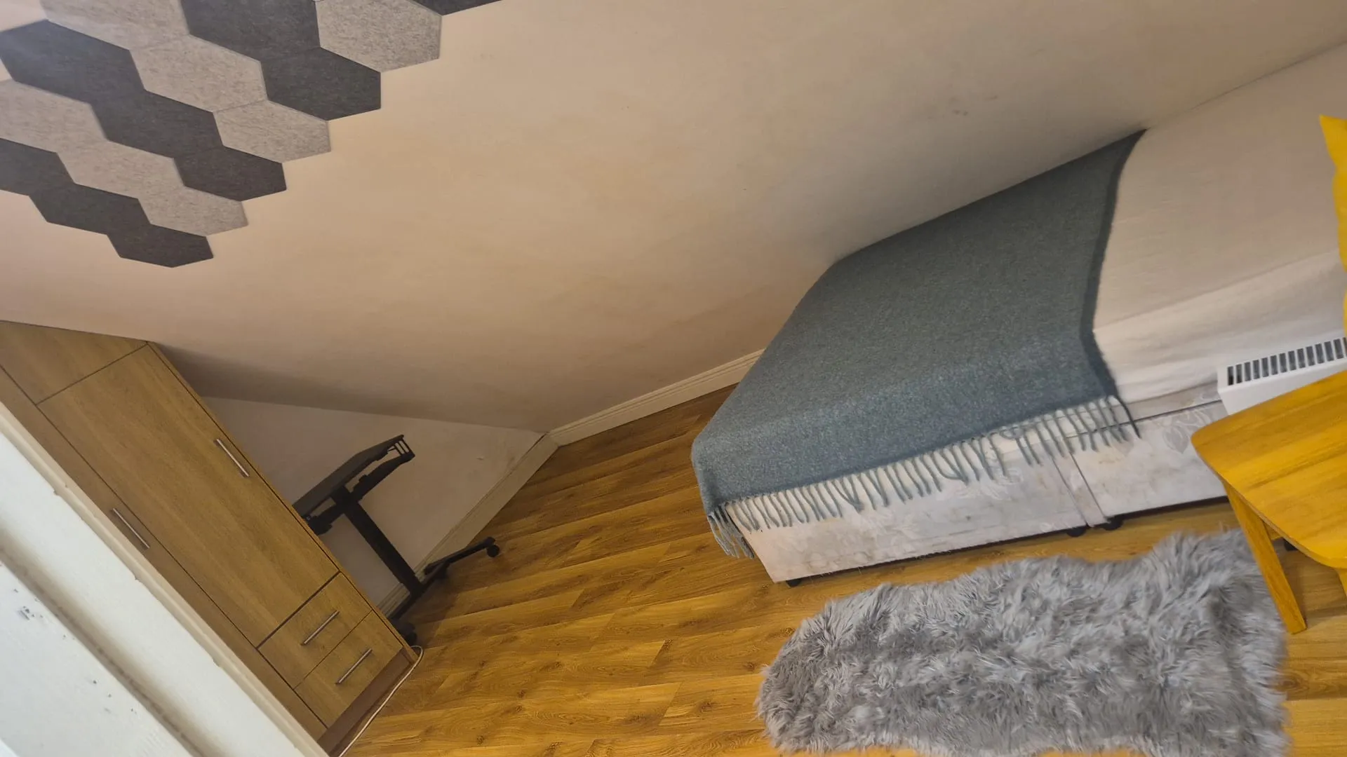Bedroom with a single bed covered by a gray throw, wooden wardrobe, office chair, small wooden side table, radiator and a gray shag rug on wood-effect floor.