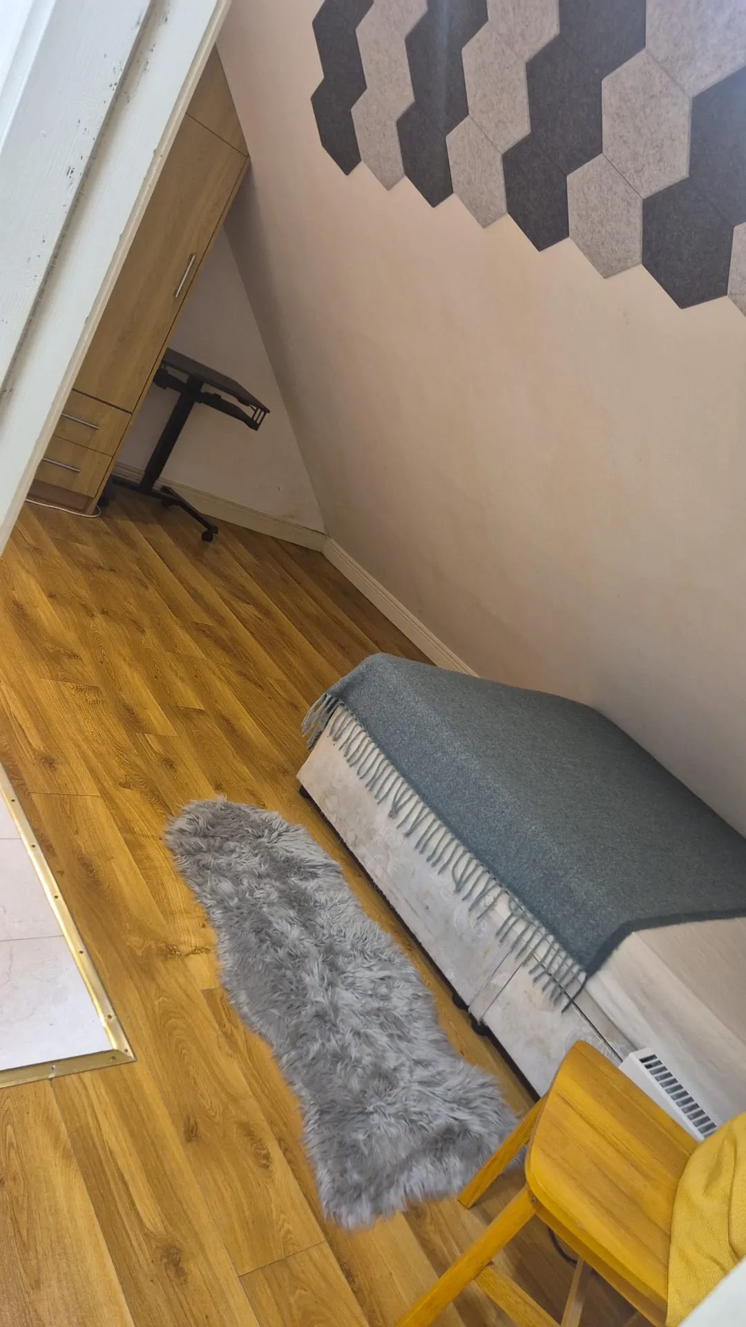 Small bedroom with a single bed covered by a gray blanket, wooden floor, long gray furry rug, wooden chair and a partial view of a desk under a sloped ceiling with hexagonal wall panels.