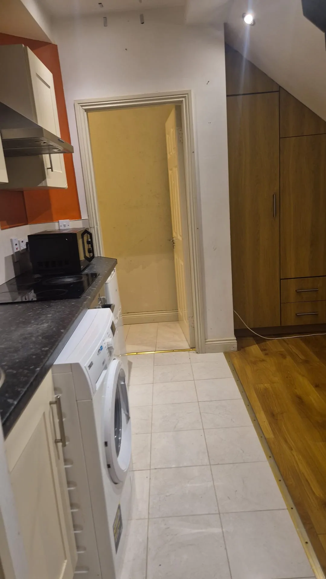 Narrow kitchen view with a washing machine, countertop with microwave and hob, overhead cabinets, and a doorway leading to another room; wood cabinets visible on the right and tiled floor transitioning to wood.