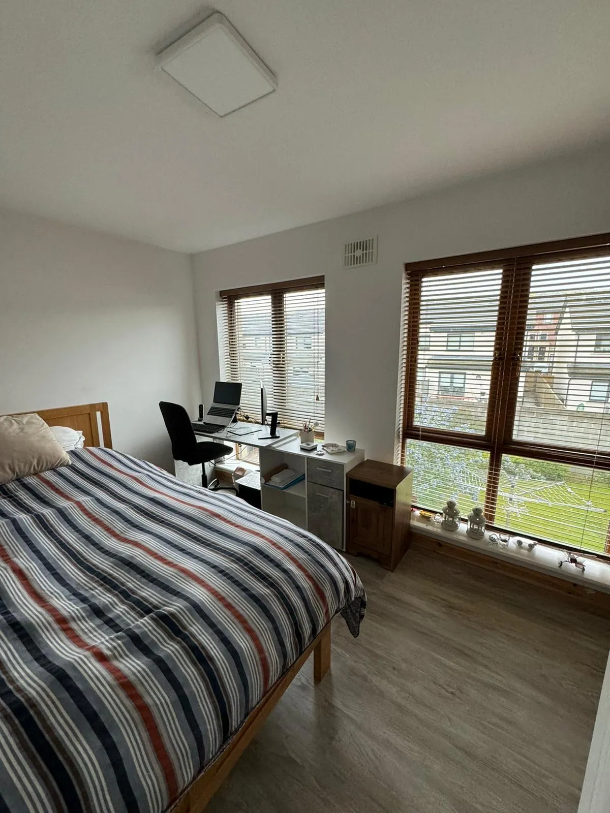 Bedroom with a double bed with striped duvet, a desk and office chair by two large windows with wooden blinds, small shelving and bedside furniture on a wood-look floor.
