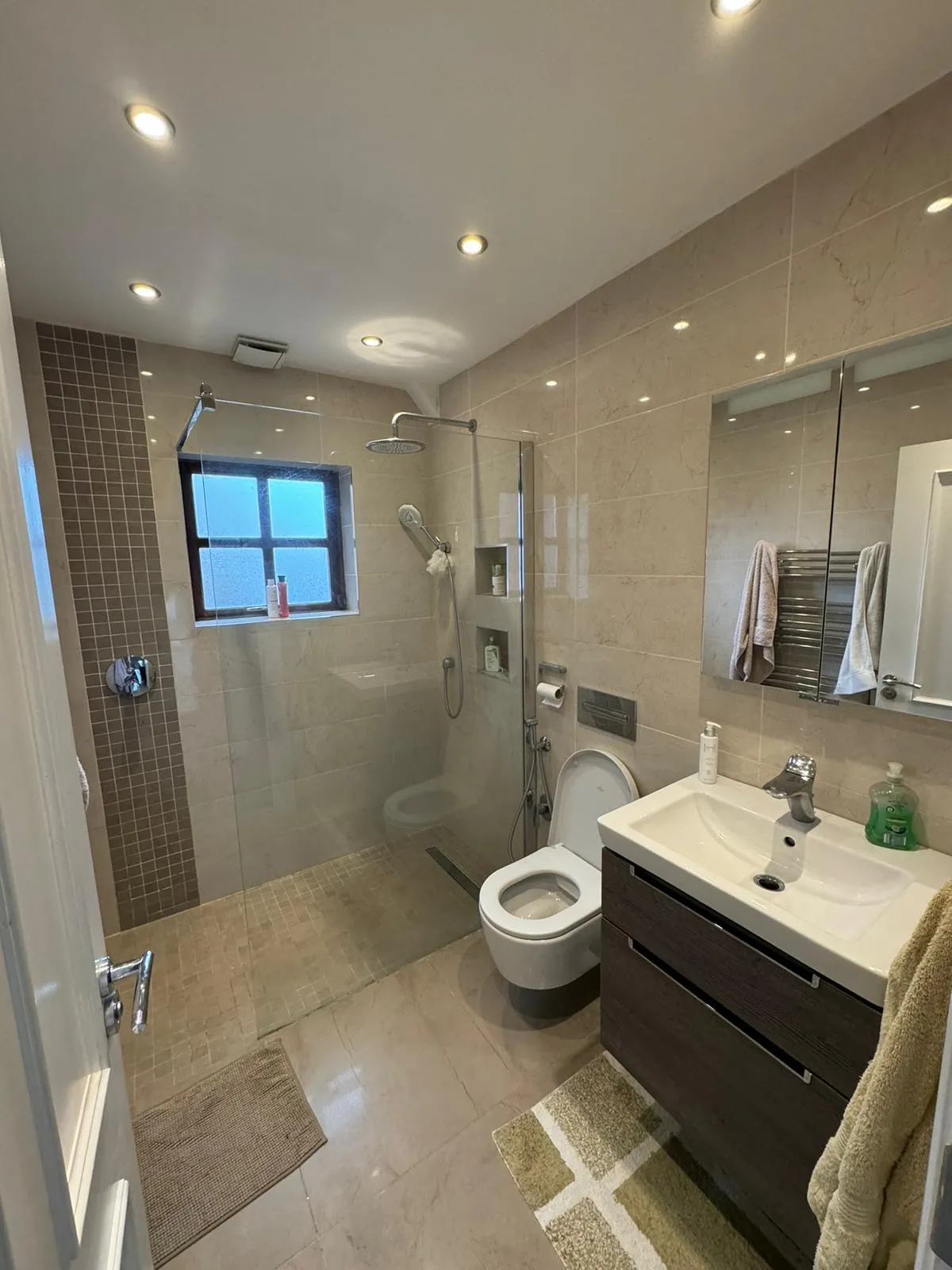Bathroom with a walk-in glass shower, wall-mounted toilet, sink with vanity and mirror, towel rack, and a small frosted window.
