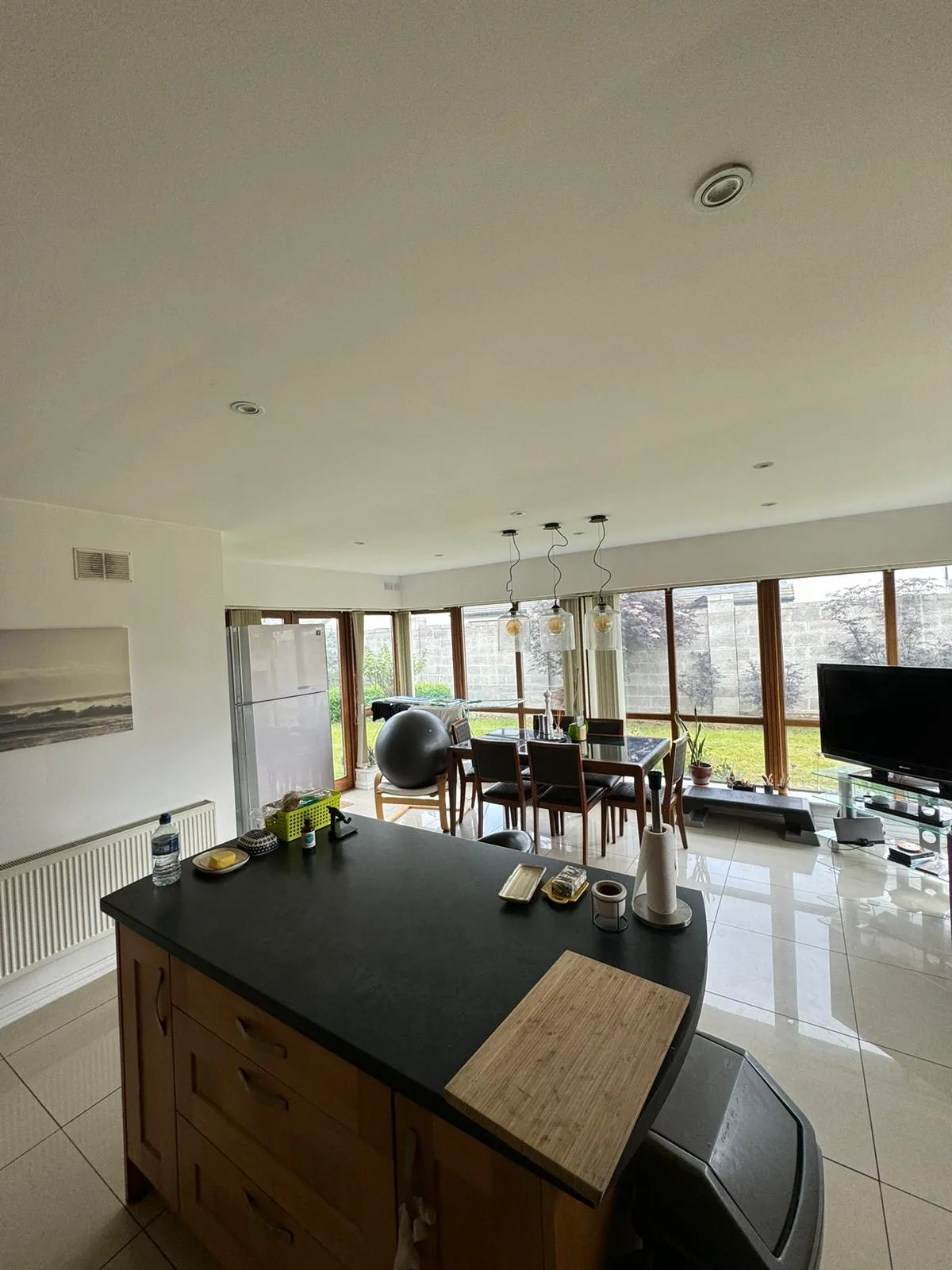 Open-plan kitchen area with a central island and countertop in the foreground, dining table with chairs and pendant lights by large floor-to-ceiling windows, refrigerator and TV visible, tiled floor and exercise ball.