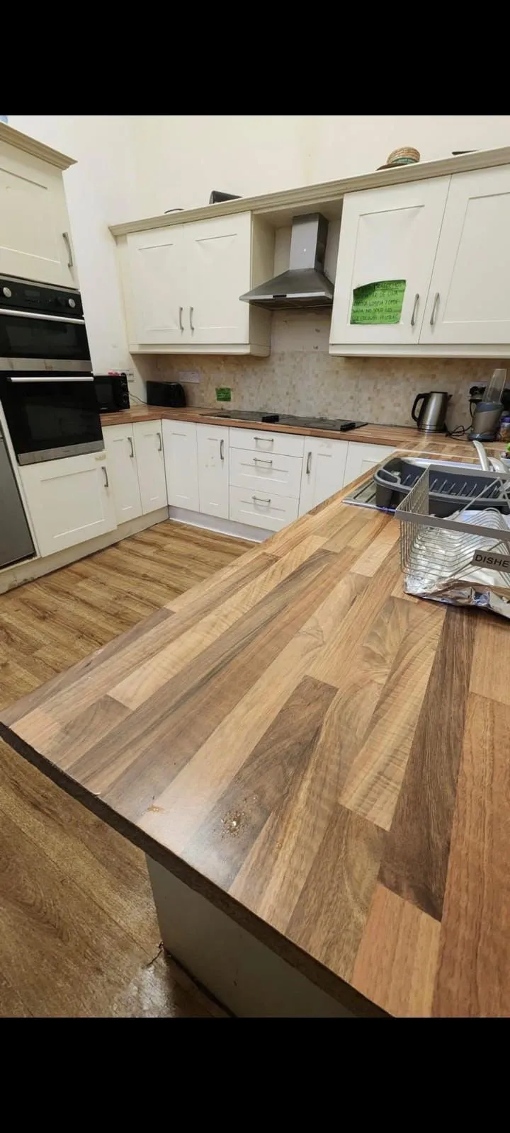 Kitchen with L-shaped counter, wooden countertops and floor, white cabinets, built-in oven, range hood, sink with dish rack and small appliances on the counter