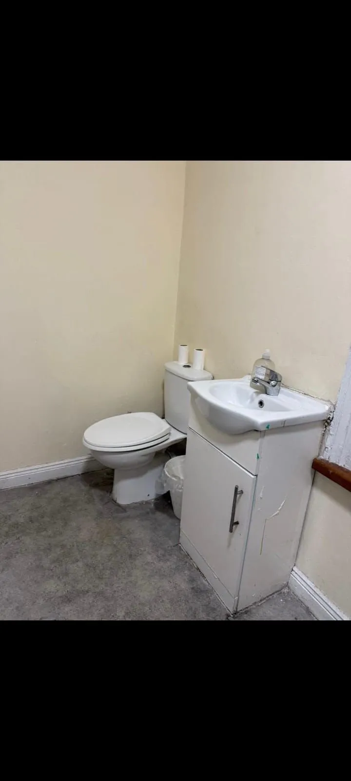Small bathroom corner with a toilet, two toilet paper rolls on the cistern, a sink set into a white vanity cabinet, and a small waste bin on the floor.