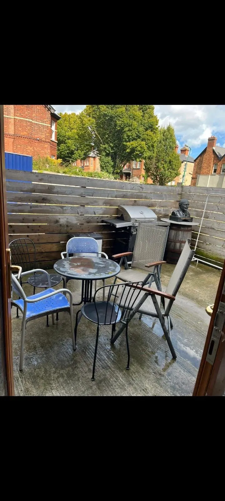 Outdoor patio with a round table, several mismatched chairs, a barbecue grill, wooden privacy fence, and concrete floor.