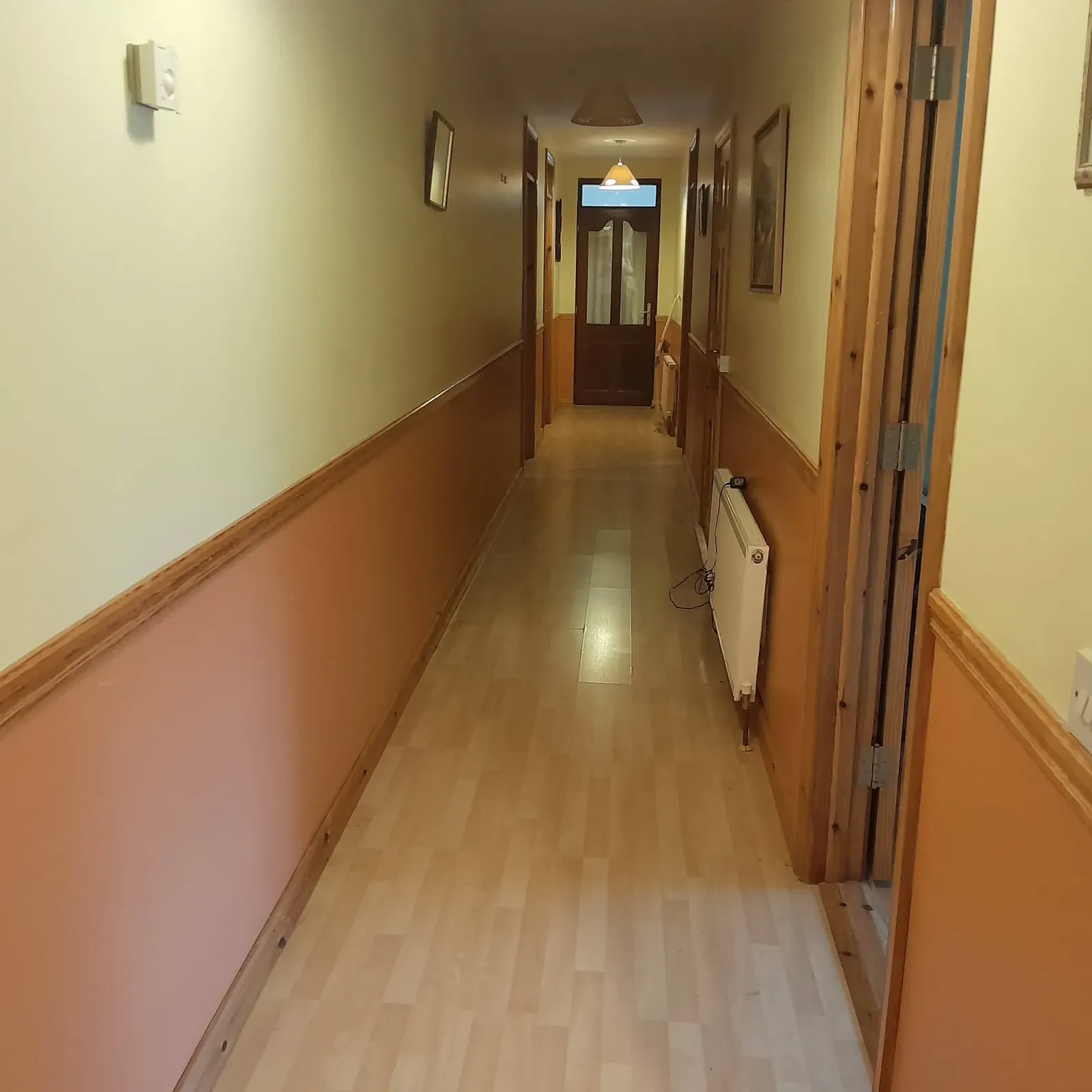 Long narrow hallway with laminate floor, wooden trim, a radiator on the right and a wooden door with a small window at the far end.
