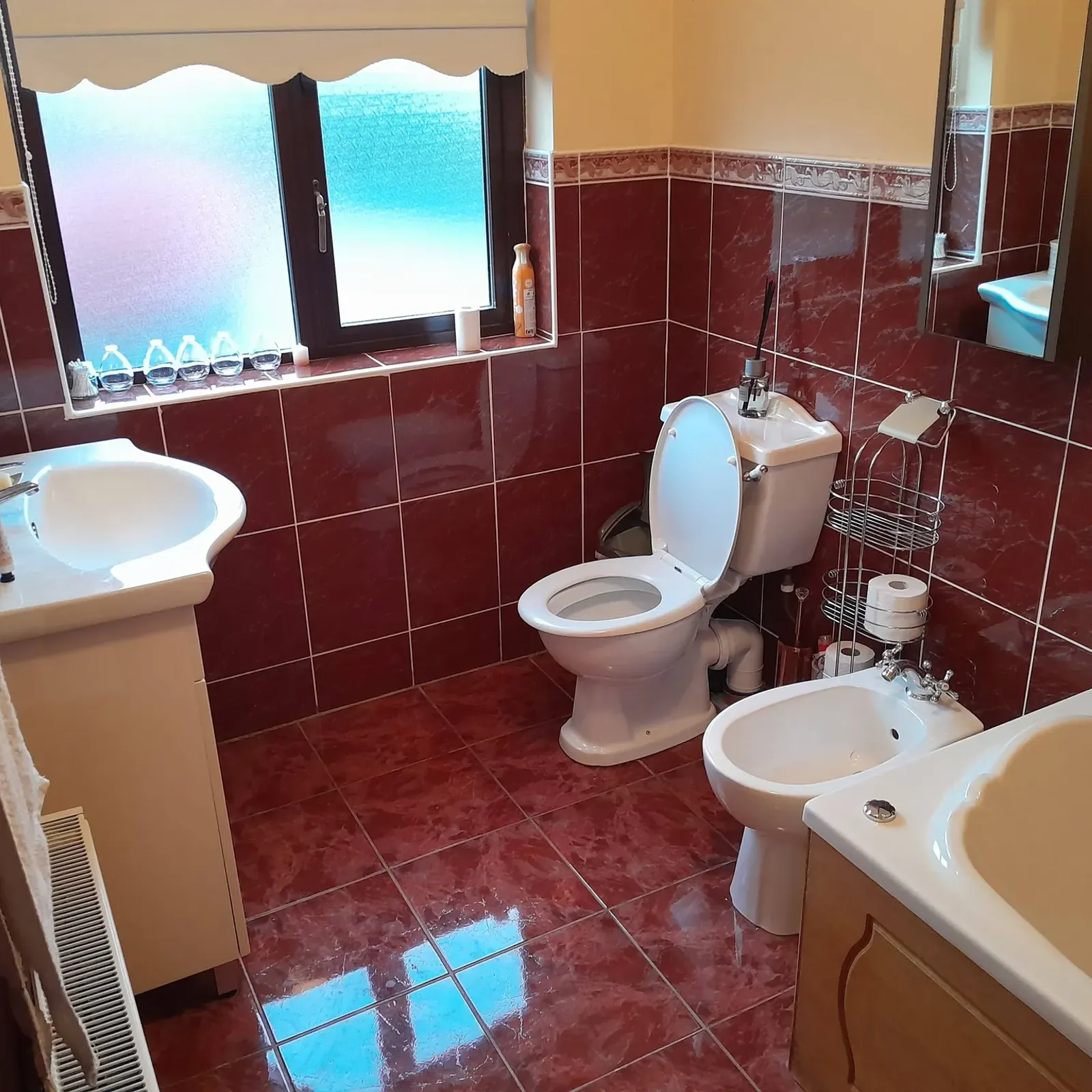 Bathroom with a toilet, bidet, sink, bathtub, wall mirror and frosted window; red tiled walls and floor.