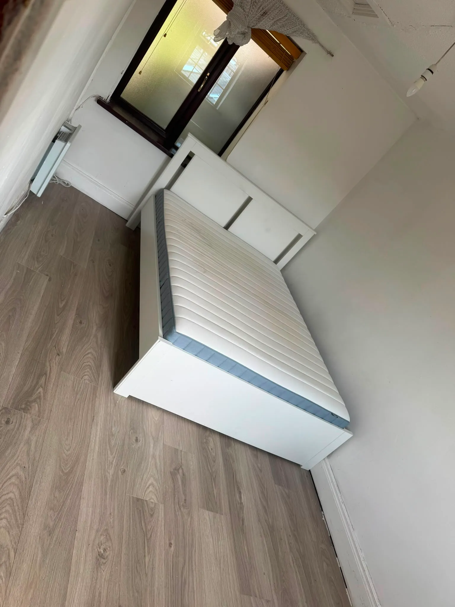Small bedroom with a white single/double bed frame and mattress, wood-look floor, frosted window with blinds, white walls and a wall-mounted heater.