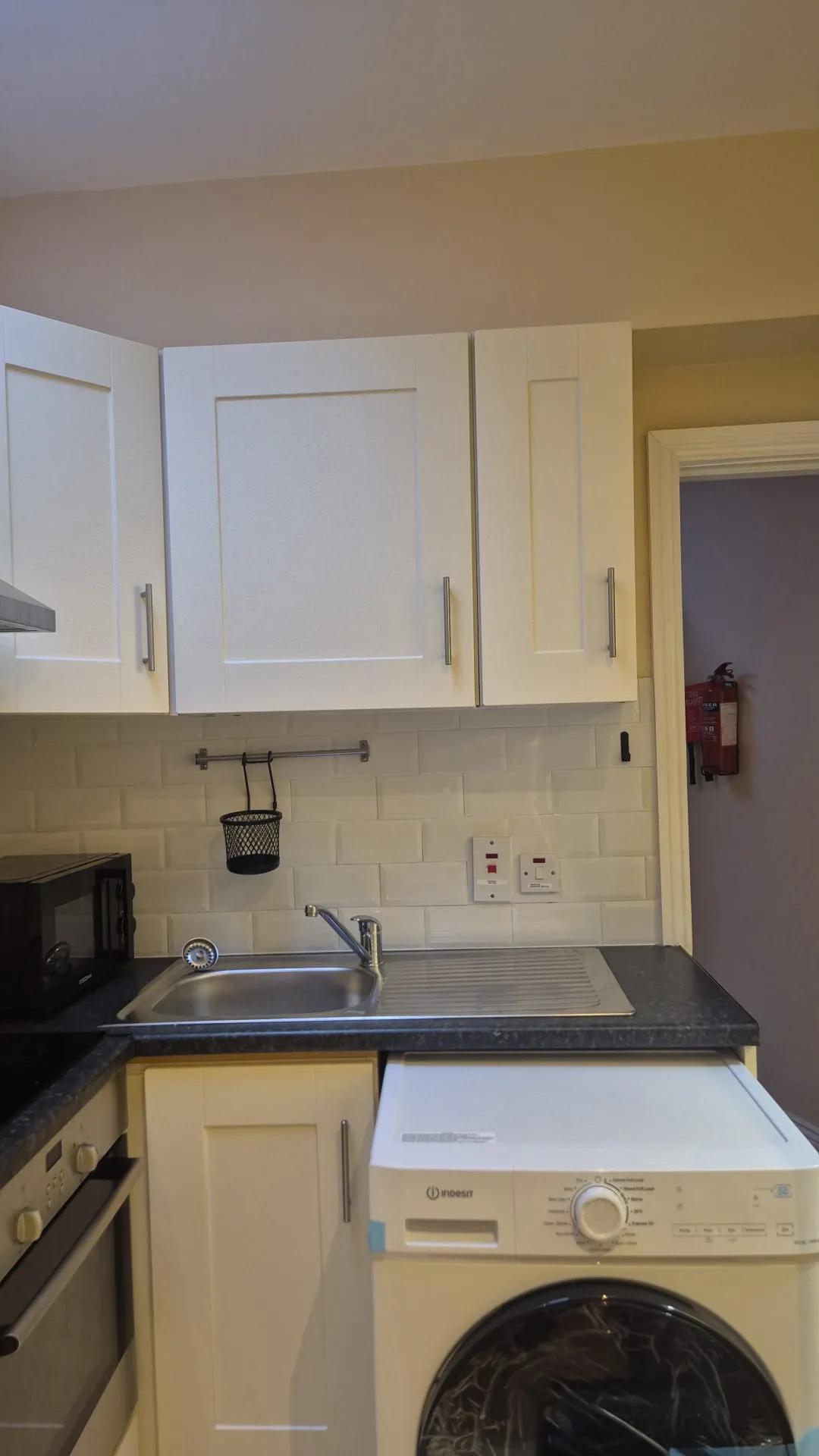 Kitchen sink and countertop with white upper cabinets, tiled backsplash, microwave and a front-loading washing machine partially visible.