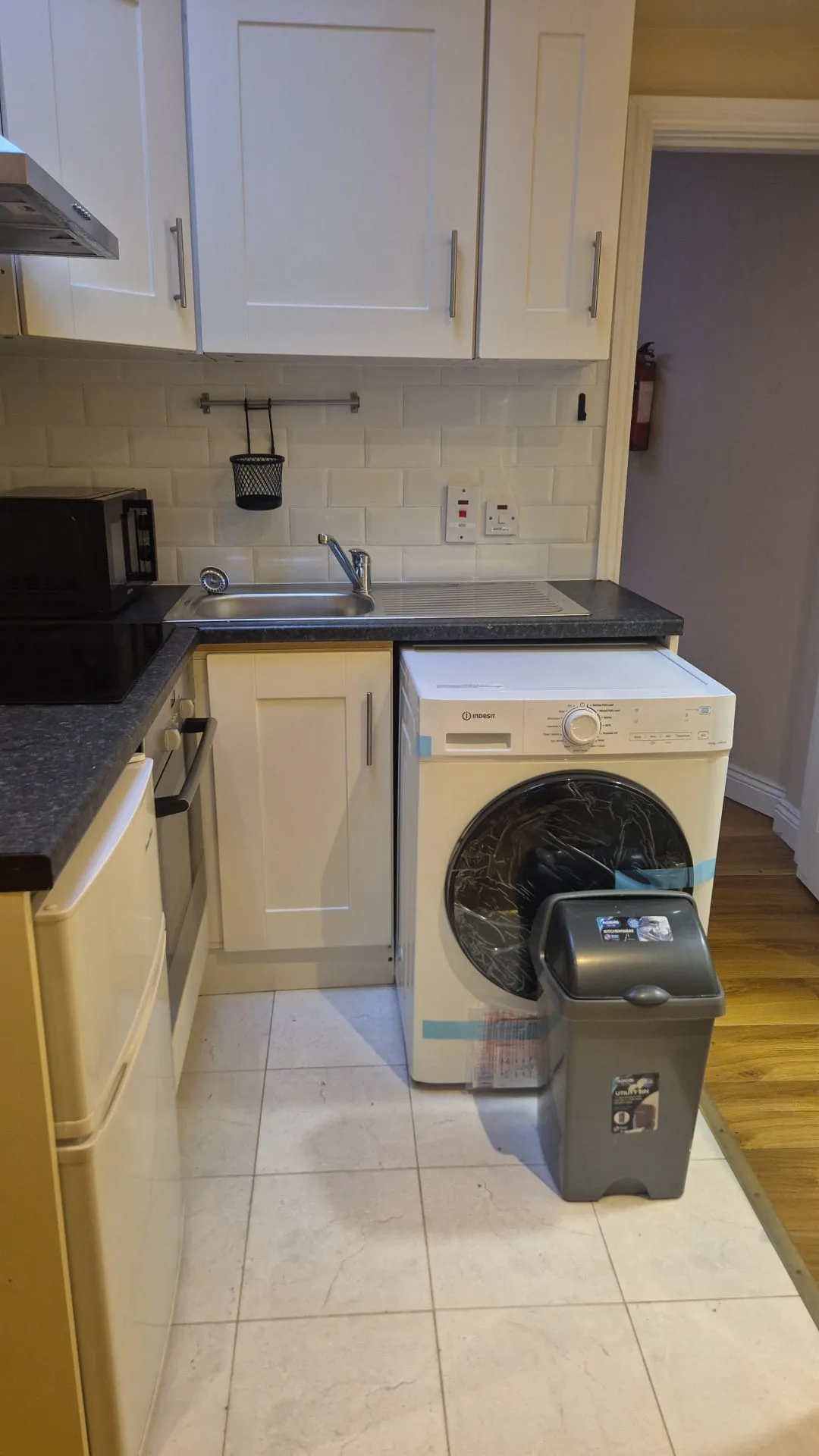Kitchen corner with white cabinets, countertop sink, microwave, front-loading washing machine and a trash bin on tiled floor