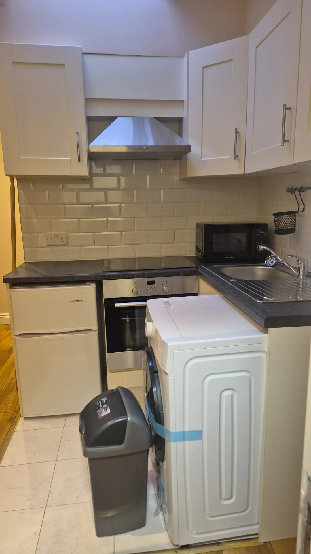 Compact kitchen with white cabinets, extractor hood above an electric oven and hob, countertop with microwave and sink, small fridge, washing machine and a trash bin in foreground.