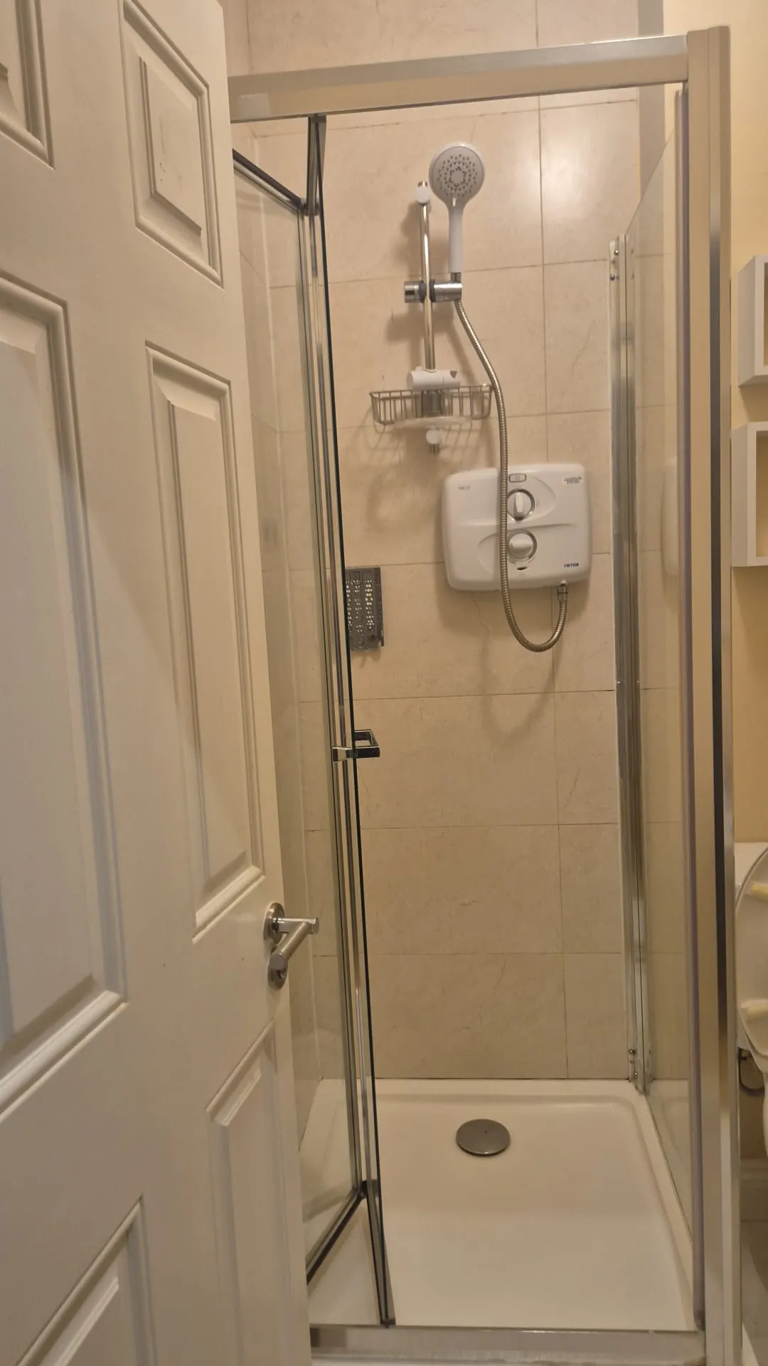 Bathroom shower stall with glass door, wall-mounted electric shower unit, handheld showerhead, and tiled walls.