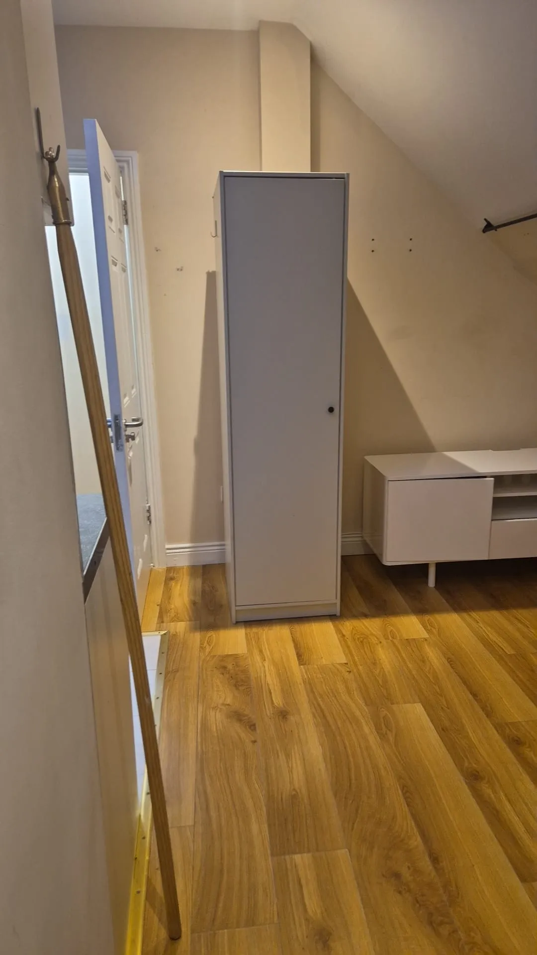 Bedroom view with a tall white wardrobe against a sloped wall, low white media cabinet, wooden floor and an open door at left.