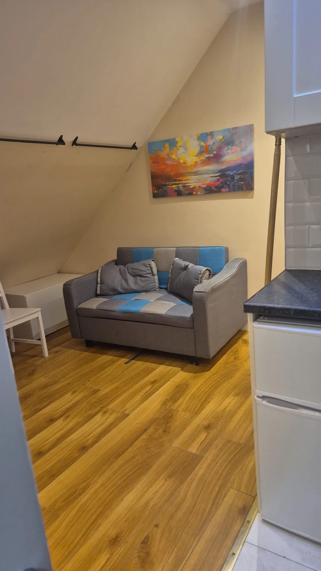 Small living area with a two-seat gray and blue sofa against a sloped wall, colorful landscape painting above, and wood-look flooring; kitchen counter and fridge visible at the right edge.