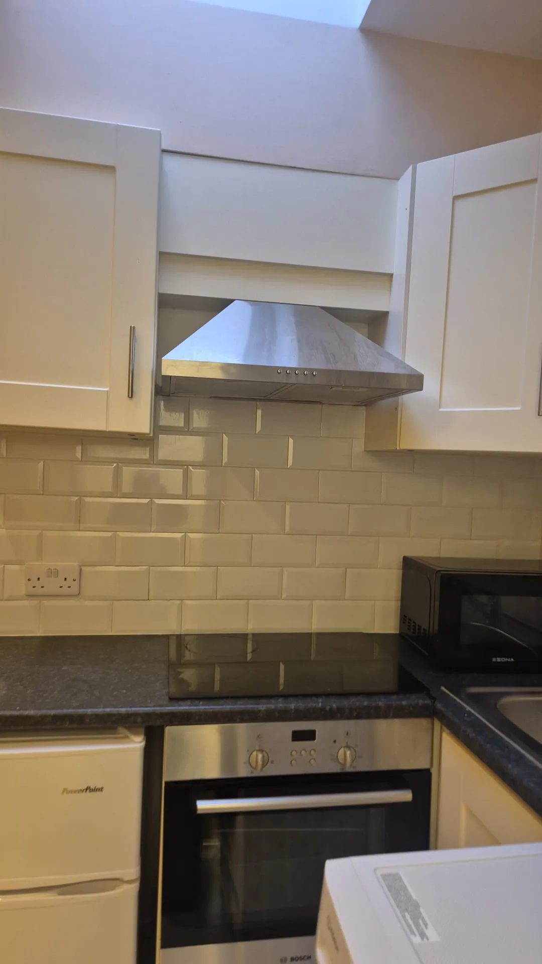 Kitchen view showing a stovetop and oven under a stainless steel range hood, white subway tile backsplash, white cabinets, black countertop and a microwave at right.