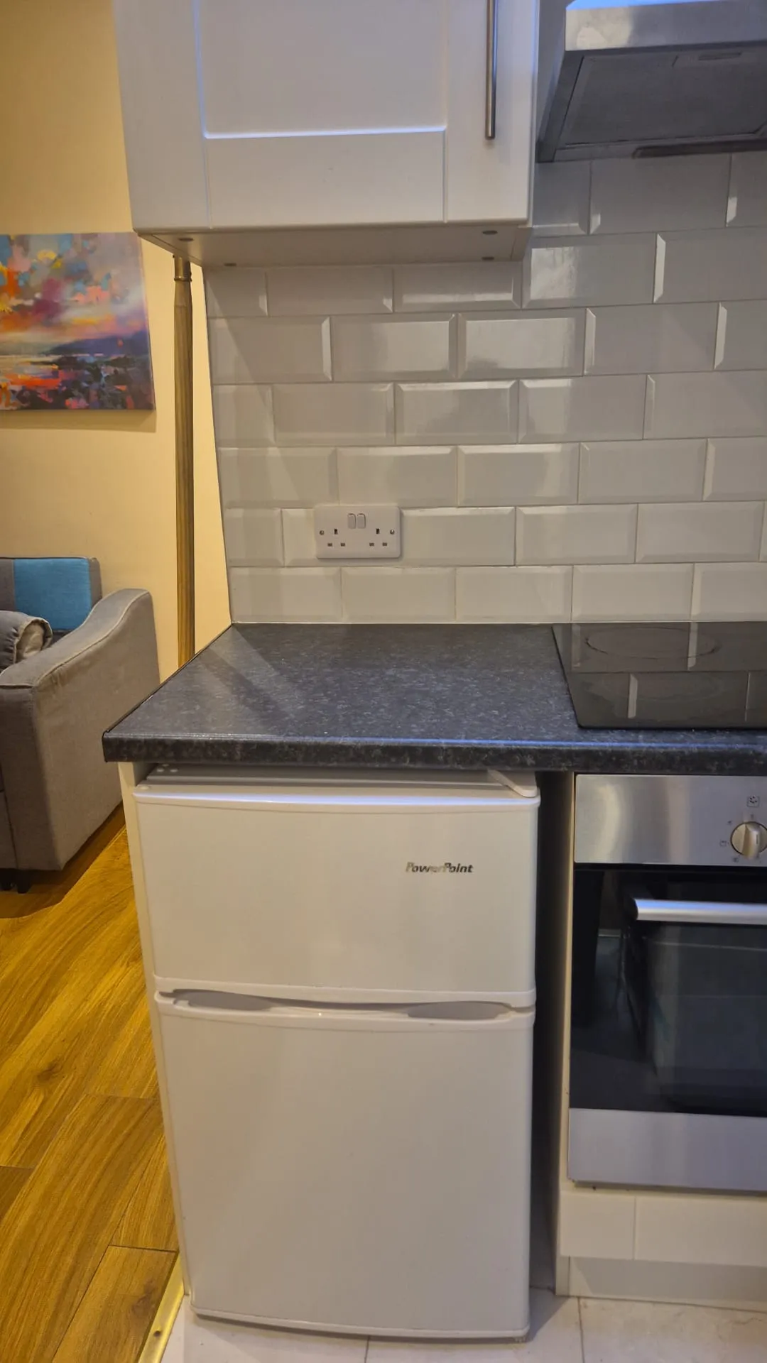 Kitchen countertop with a small white fridge under a dark countertop, tiled backsplash, wall power outlet, and part of an oven and cabinet visible.