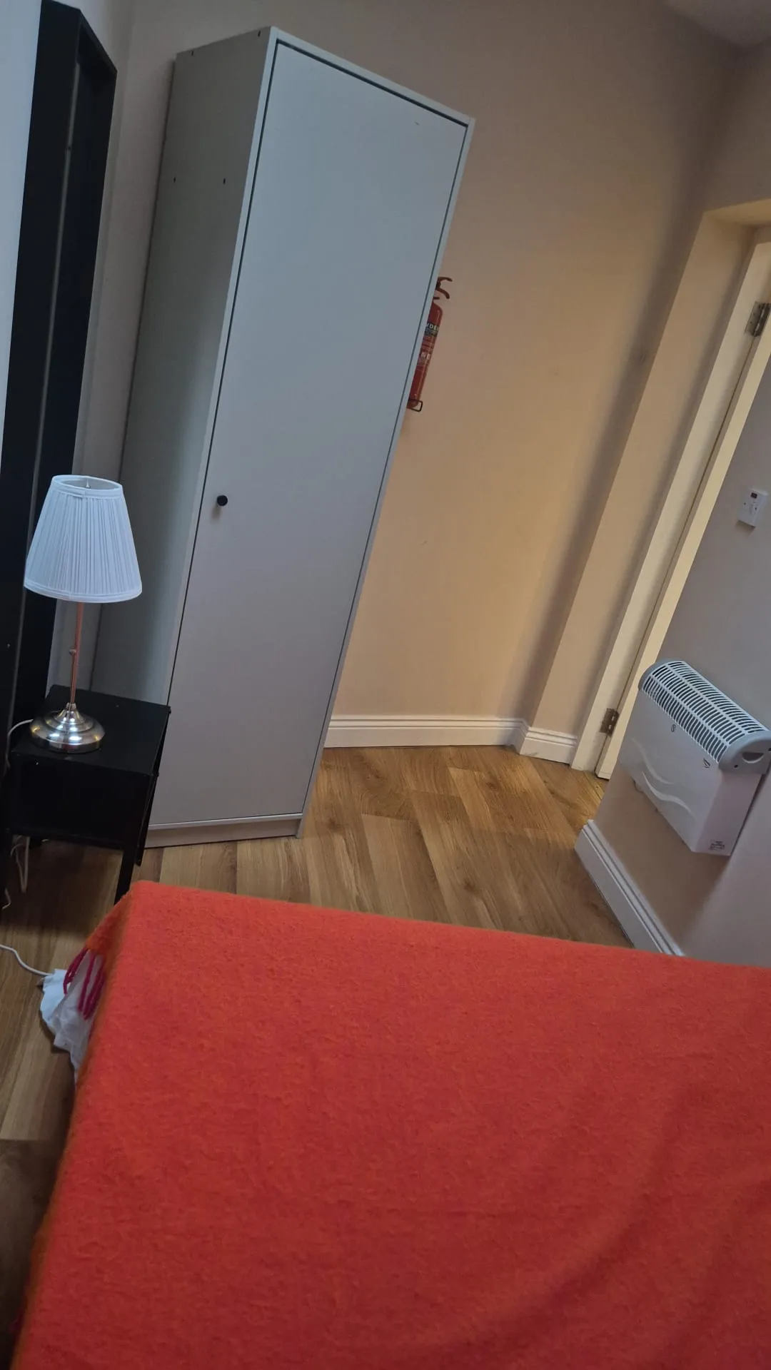 Bedroom corner with a red blanket on a bed, a tall white wardrobe, small black bedside table with a lamp, and a wall-mounted heater.