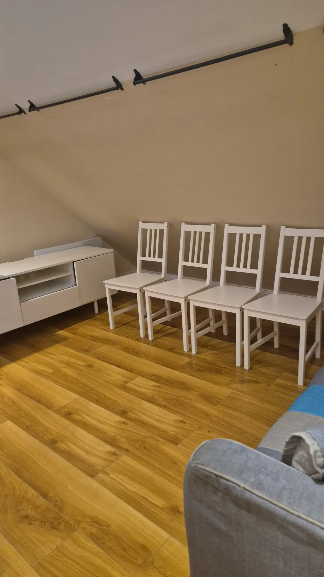 Living room with four white chairs lined against a sloped wall, a low white media console, wood-look flooring and part of a gray sofa visible.