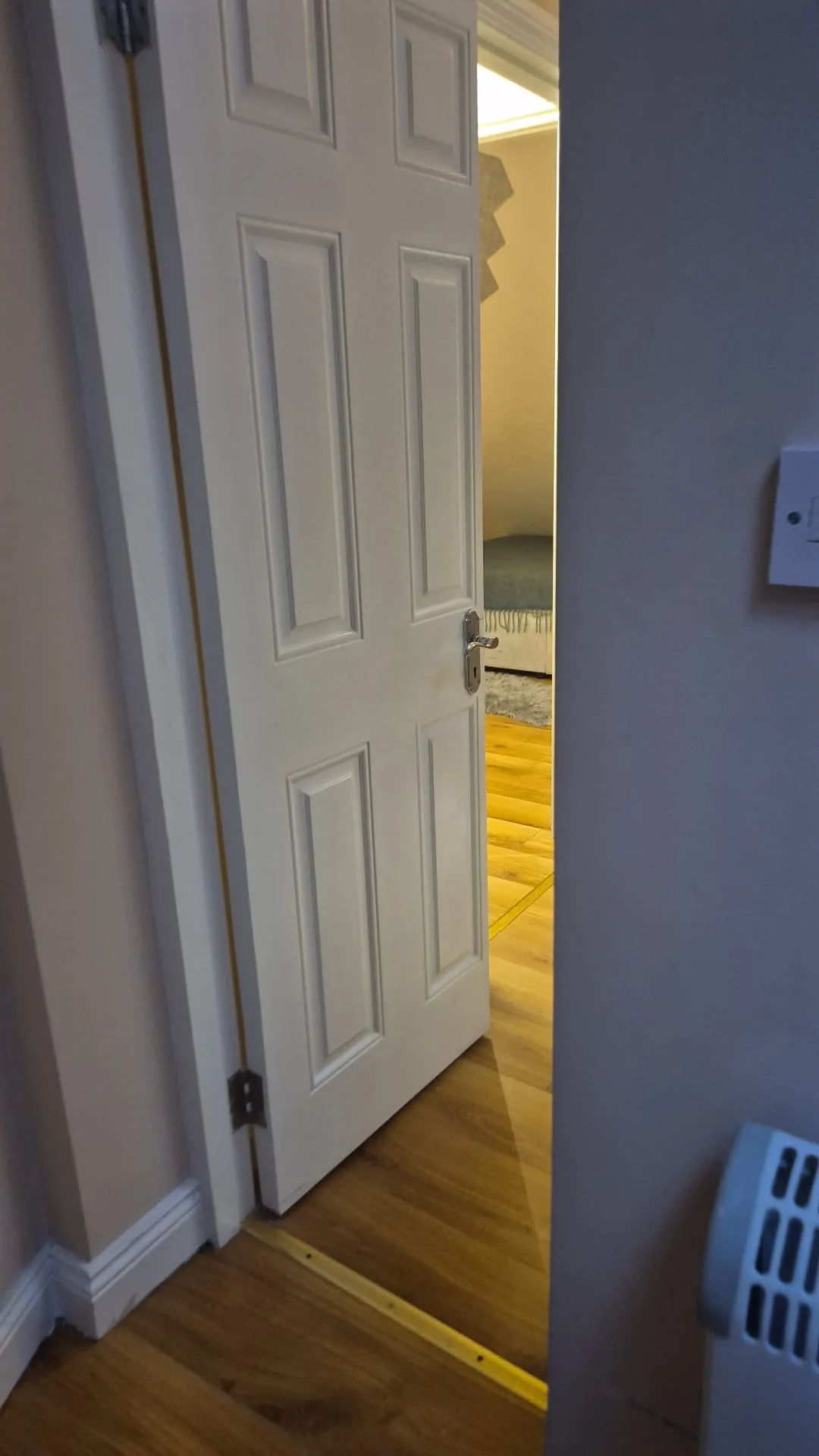 Partially open interior door in a hallway showing wooden floor and a small view into a bedroom with a bed and rug.