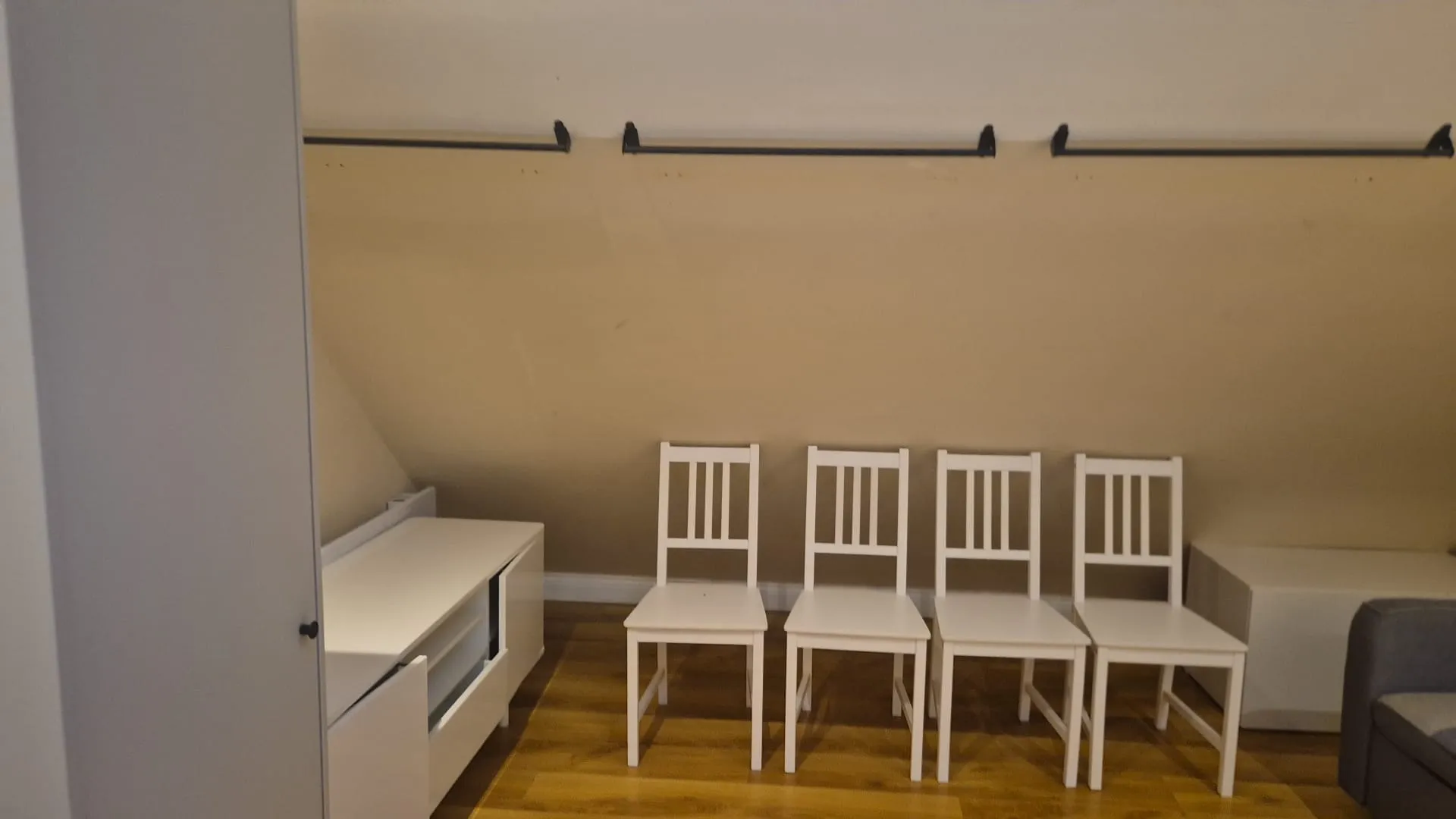 Room with a sloped ceiling, four white wooden chairs in a row, a low white cabinet and a wardrobe at left