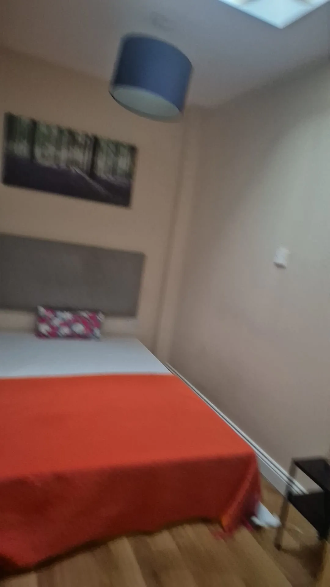 Bedroom with a bed covered by an orange blanket, a headboard, wall art, a pendant ceiling light and a small bedside table.