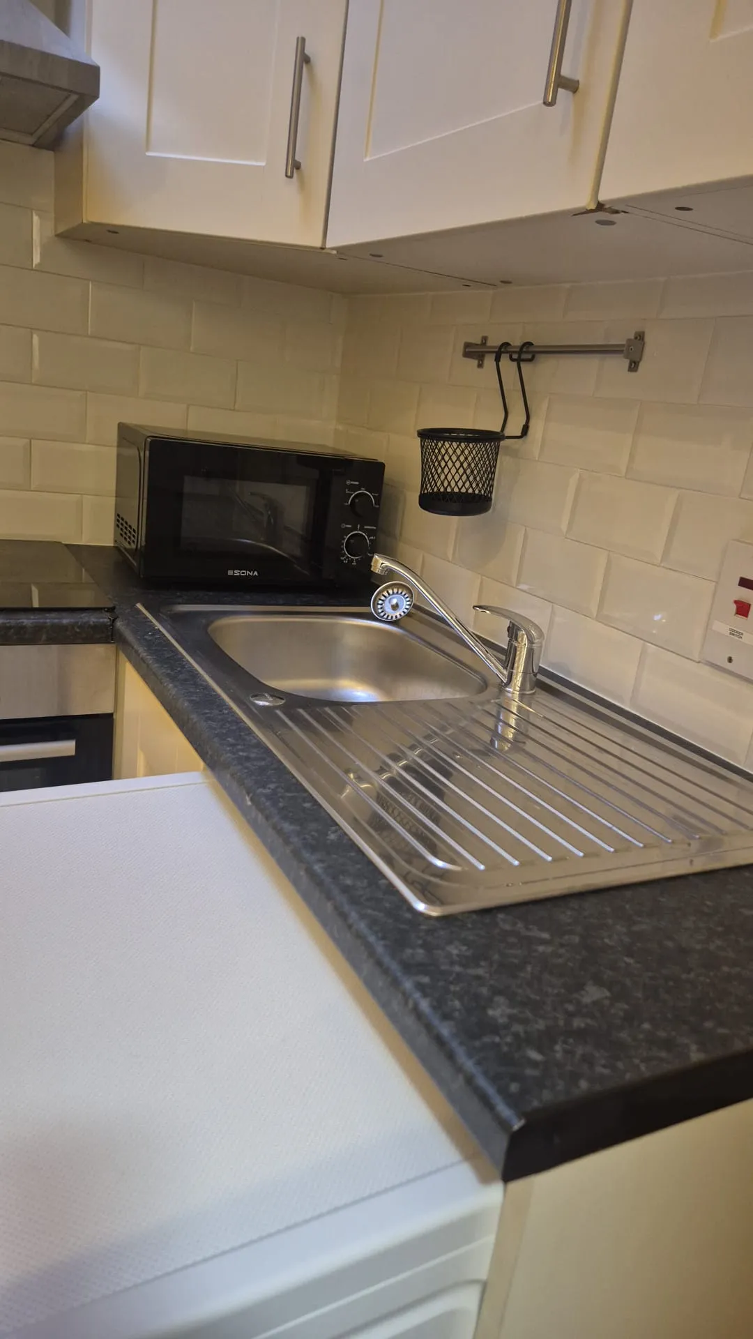 Kitchen countertop with a stainless steel sink and drainer, single-handle faucet, microwave on the counter, white cabinets and tiled backsplash with a small hanging basket.