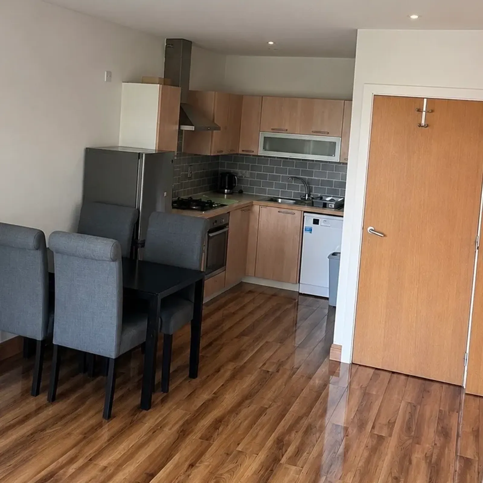 Open-plan kitchen with wooden cabinets, tiled backsplash, stainless refrigerator, oven and sink, and a dining table with four gray chairs on wood-look flooring.