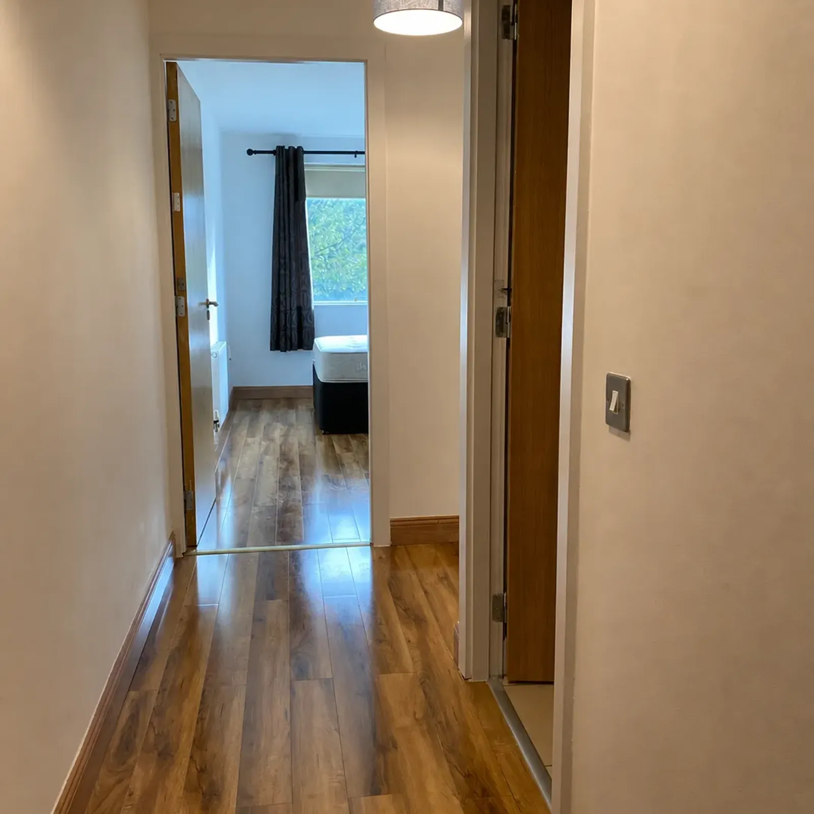 Hallway with wood-look floor, overhead pendant light and an open doorway leading to a bedroom with a window and curtains