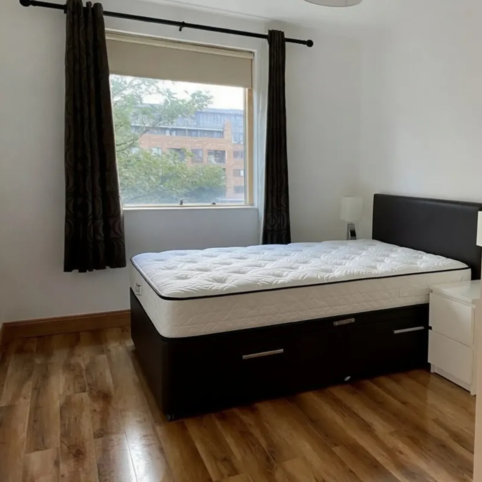 Bedroom with a double bed on a black upholstered divan base, white mattress, bedside table with lamp, window with roller blind and dark curtains, and wood-look floor.
