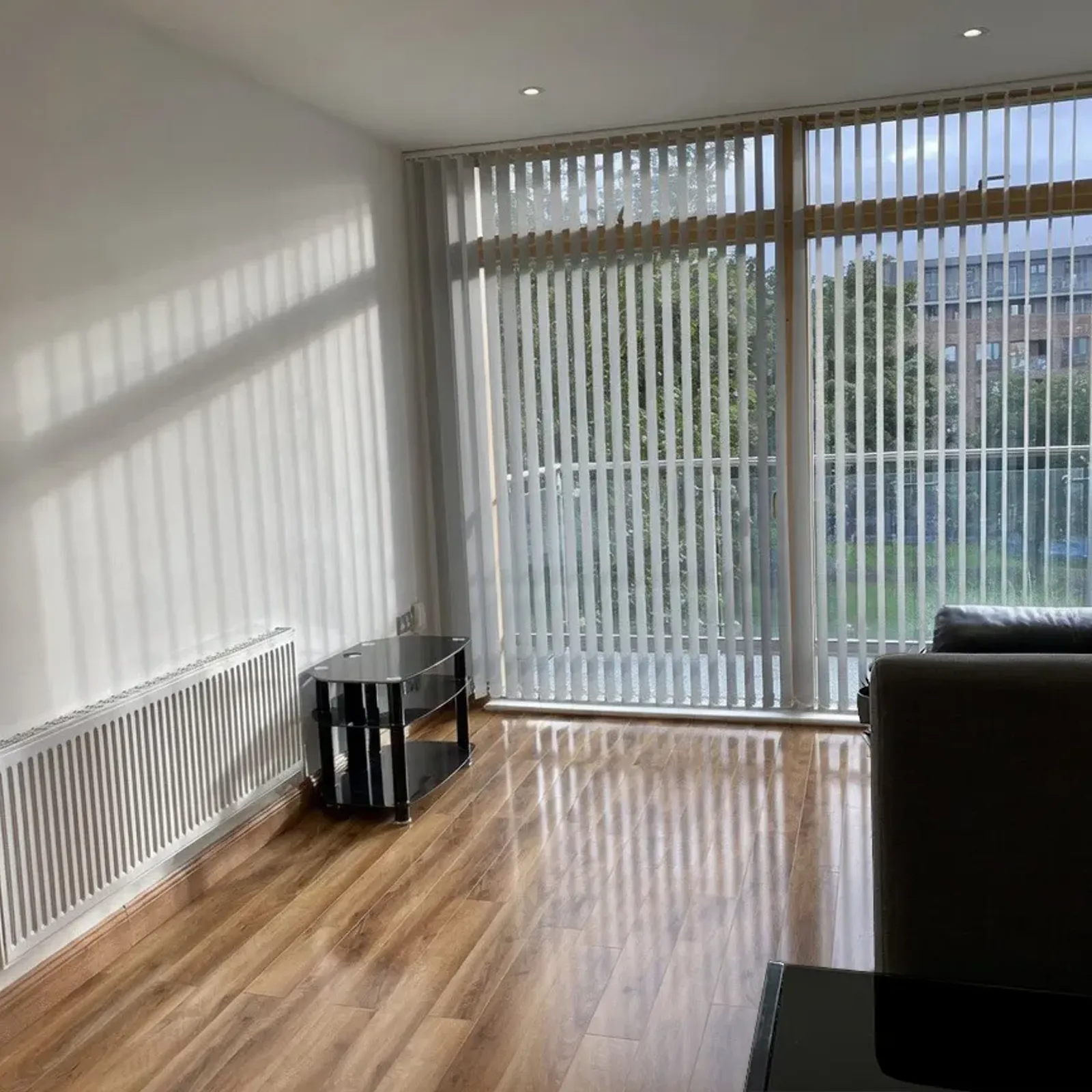 Living room with large floor-to-ceiling windows with vertical blinds, hardwood floor, radiator, small glass side table and a dark sofa partially visible