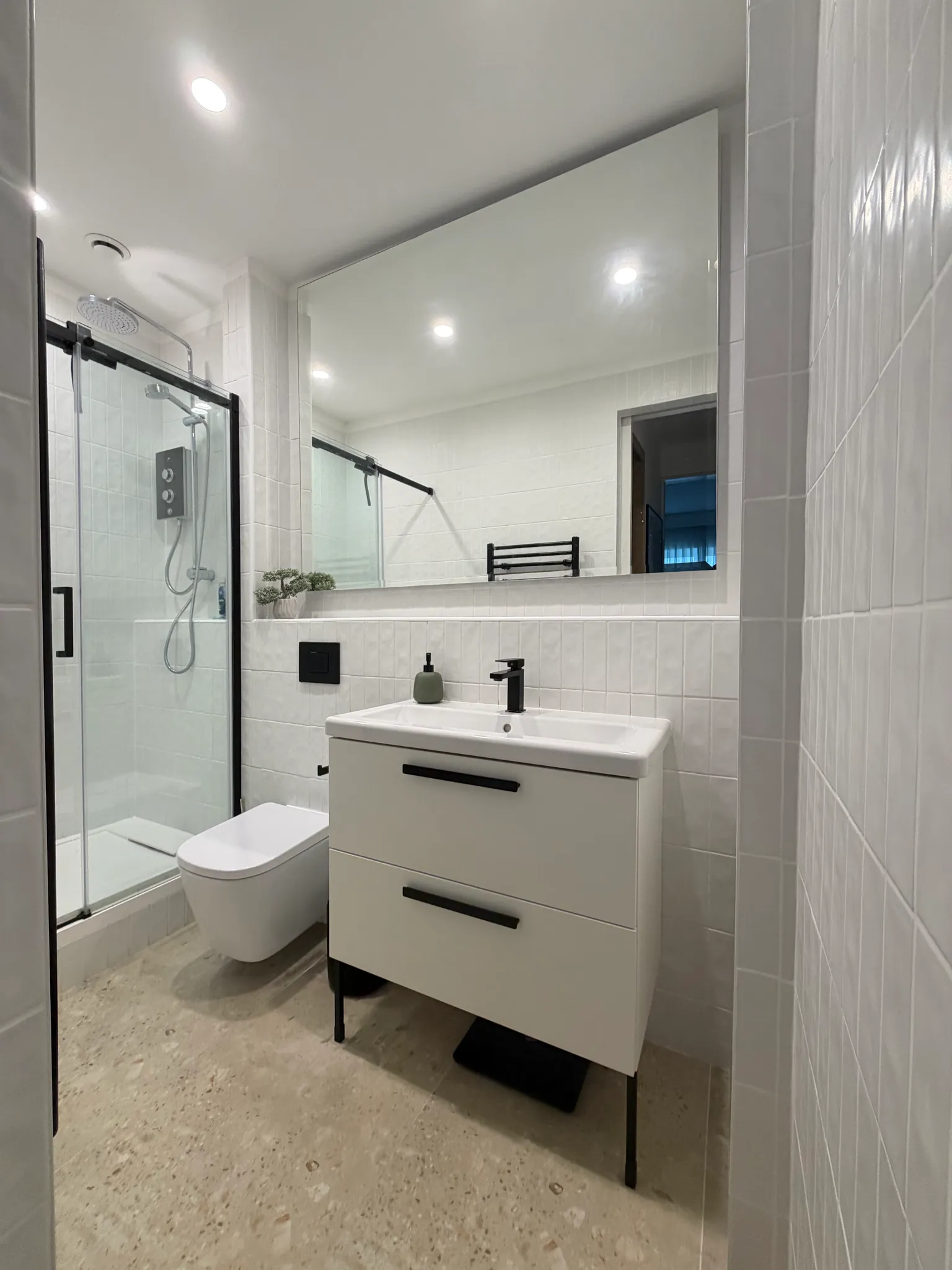Bathroom with a glass shower enclosure, wall-mounted toilet, white vanity with sink and mirror, and tiled walls.
