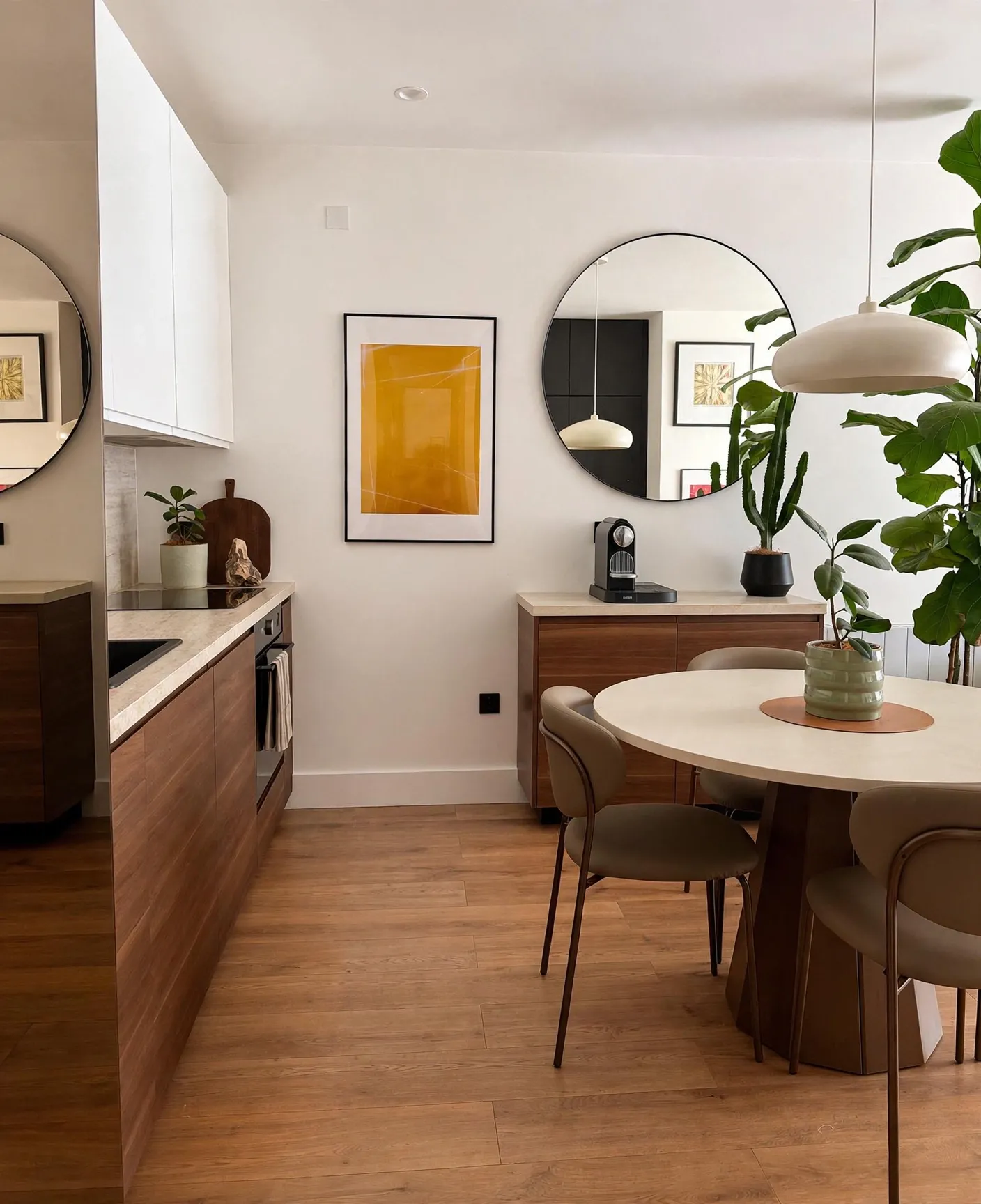 Dining area and kitchen corner with a round mirror, pendant light, round dining table with chairs, wooden cabinets, countertop with a coffee machine, and several potted plants.