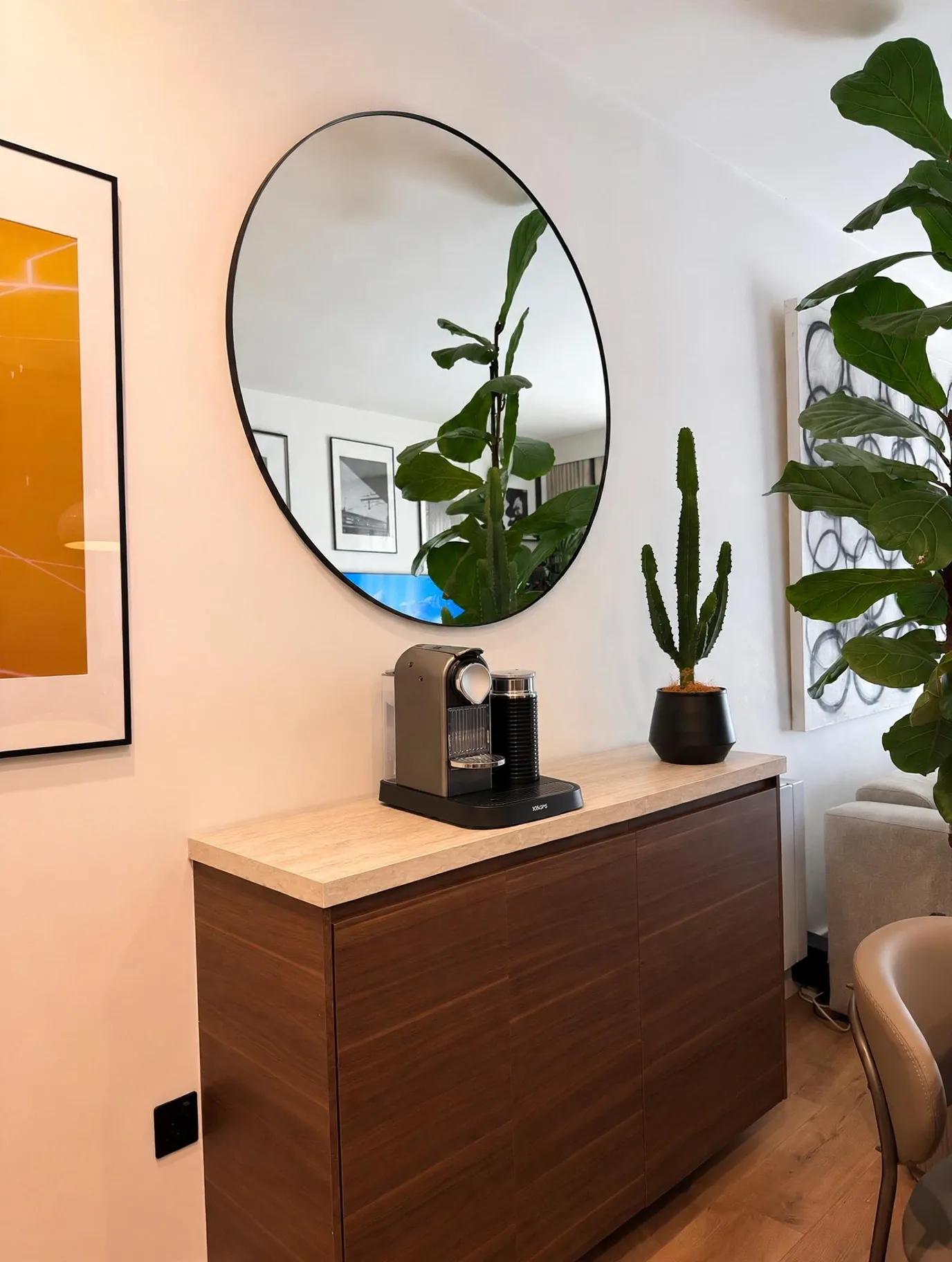Sideboard with a coffee machine and potted cactus under a round wall mirror reflecting plants and framed art.