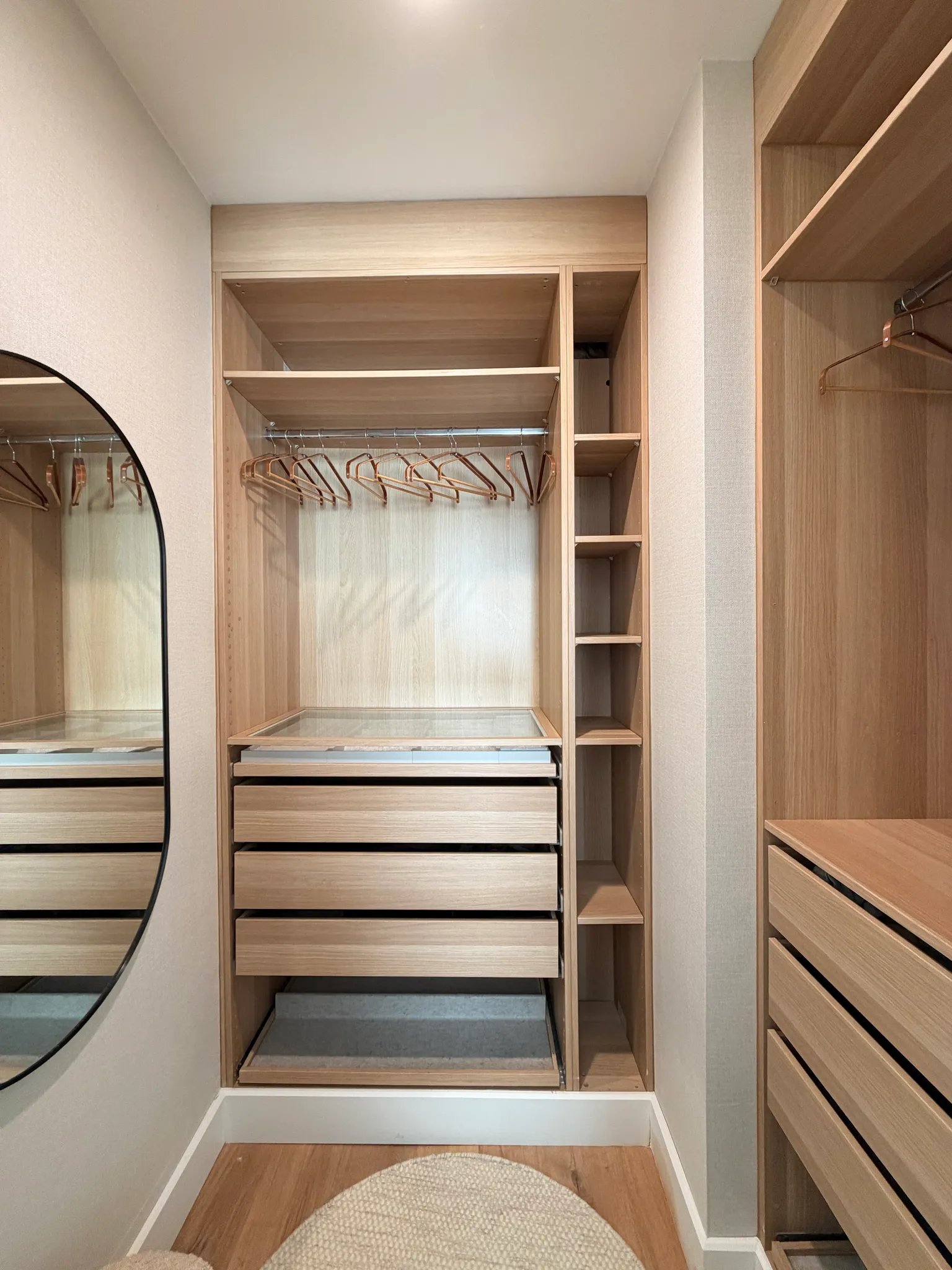 Walk-in closet with wooden built-in shelving, drawers, hanging rod with wooden hangers and a partial oval mirror on the left.