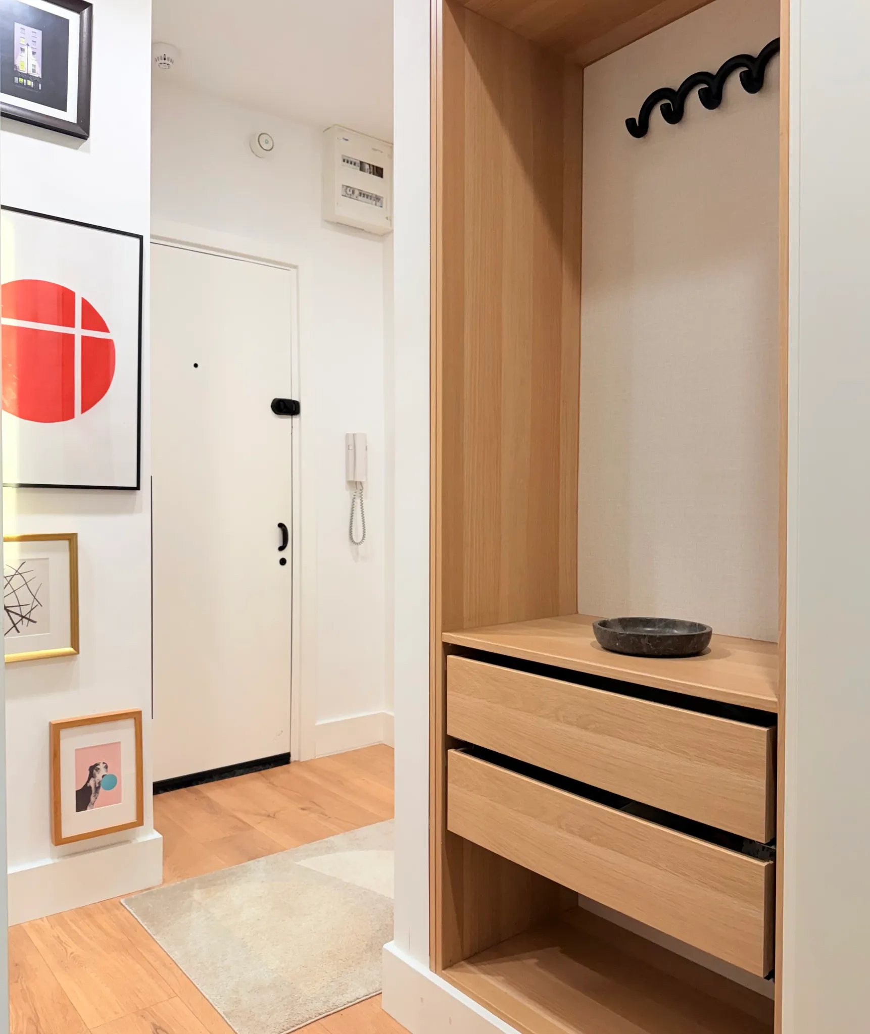 Entry hallway with a white front door, wall intercom, framed artwork, a wooden built-in closet with drawers and coat hooks, and a small bowl on the shelf.