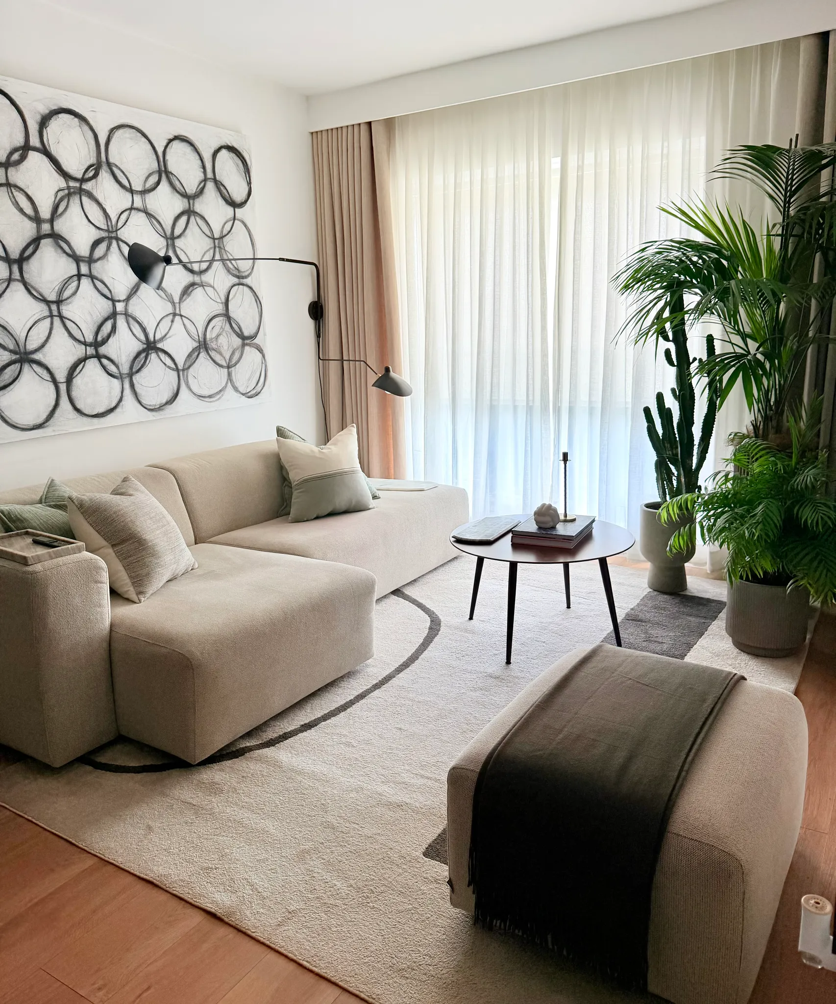 Living room with a beige sectional sofa, throw pillows, round coffee table, large sheer curtains, wall lamp, abstract circular wall art and potted plants.