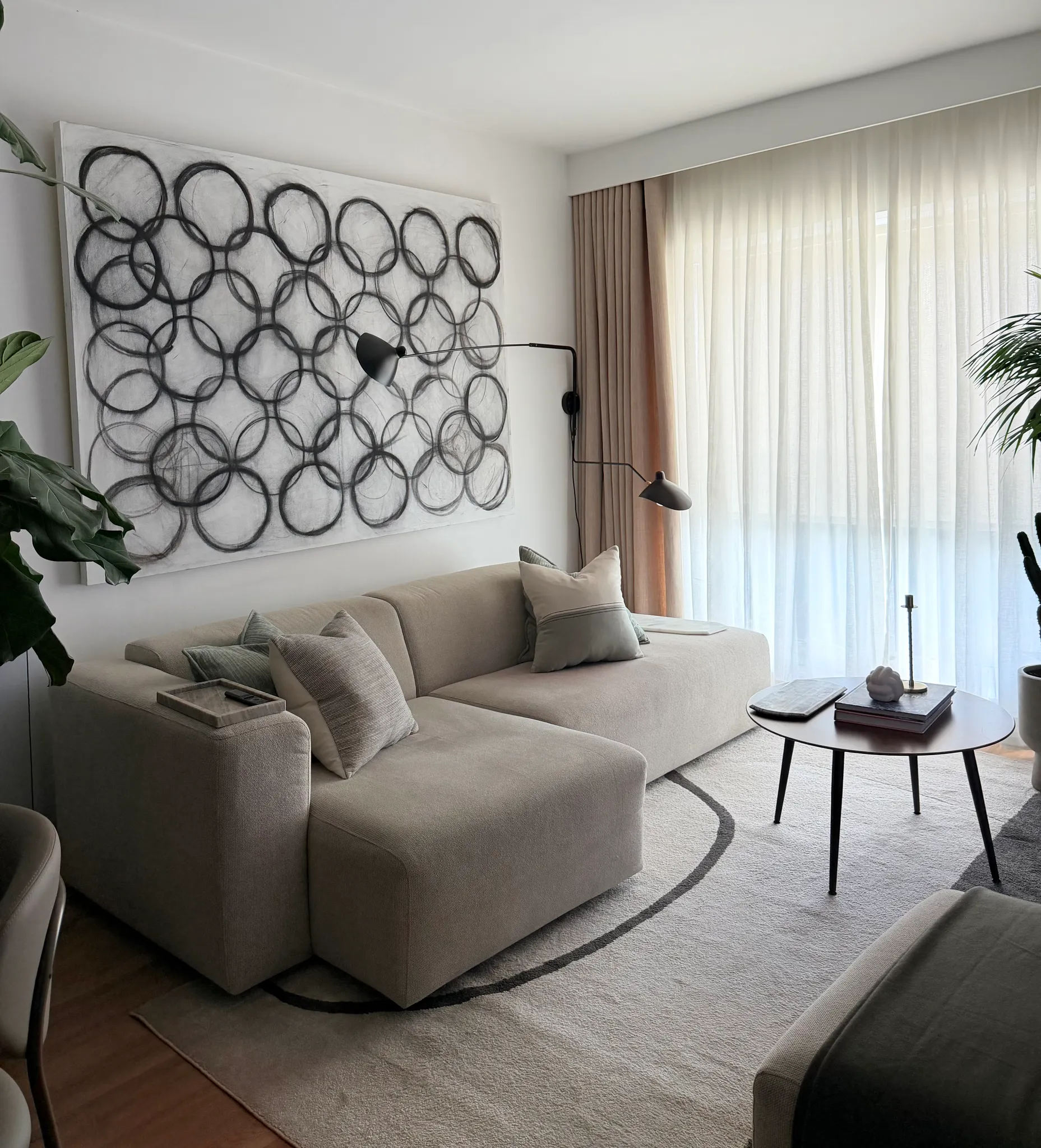 Living room with a beige sectional sofa, decorative pillows, round coffee table, large abstract wall art, floor lamp, sheer curtains and a potted plant.