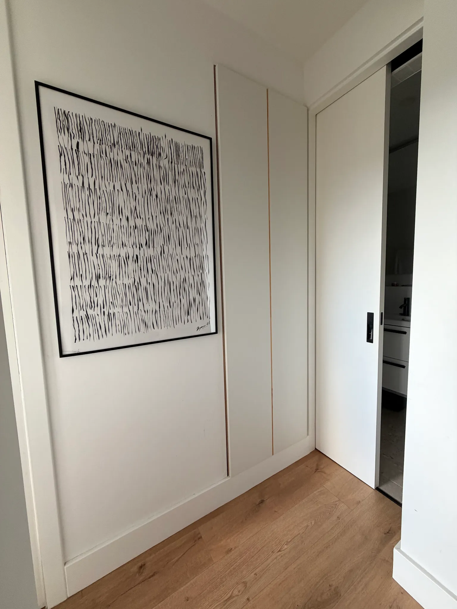 Hallway with framed abstract artwork on the wall, built-in white closet panels and a partially open white sliding door revealing an adjacent room.
