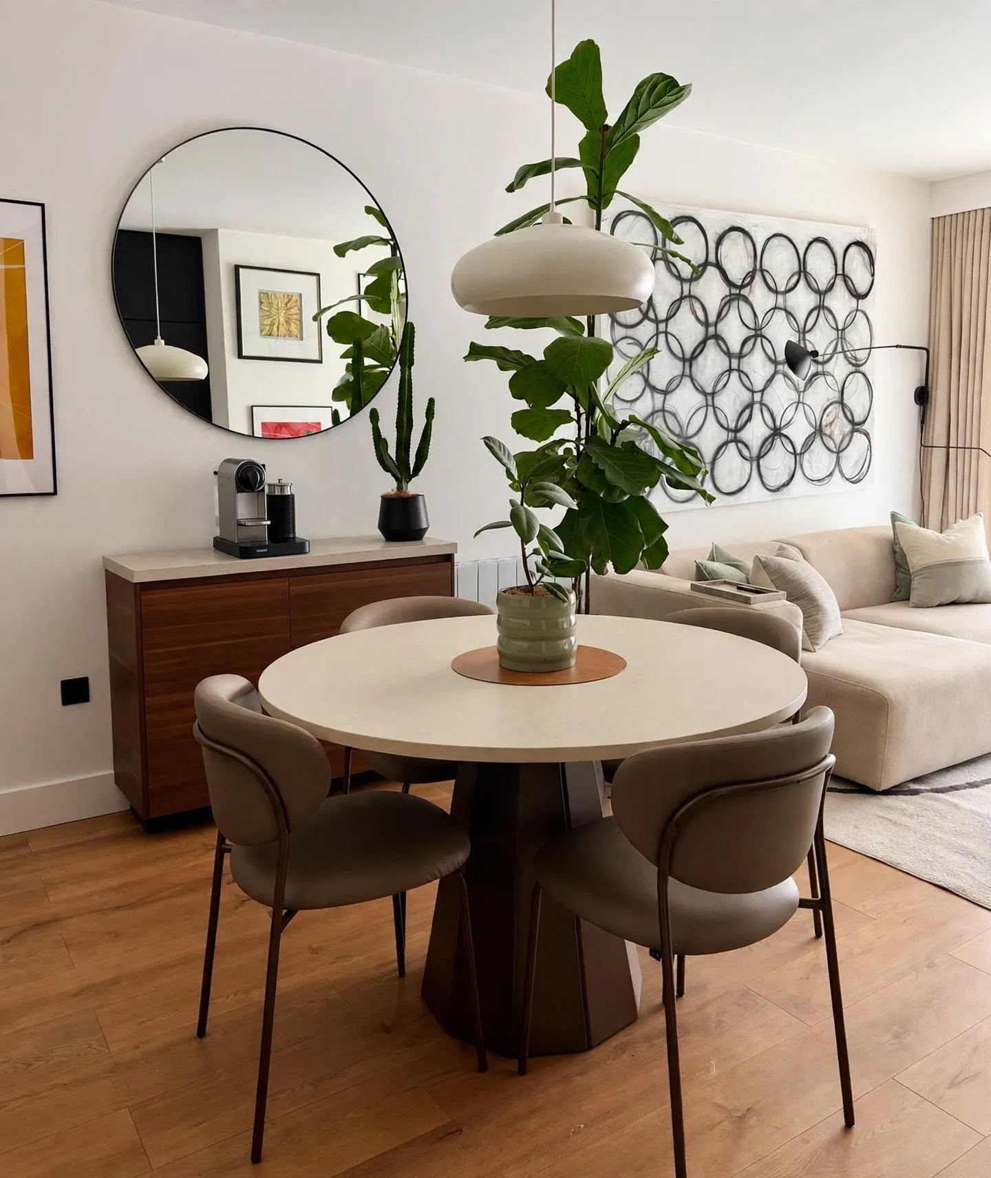 Dining area with a round table, four chairs, pendant lamp, tall potted plant, sideboard with coffee machine, and a sofa with wall art in the background