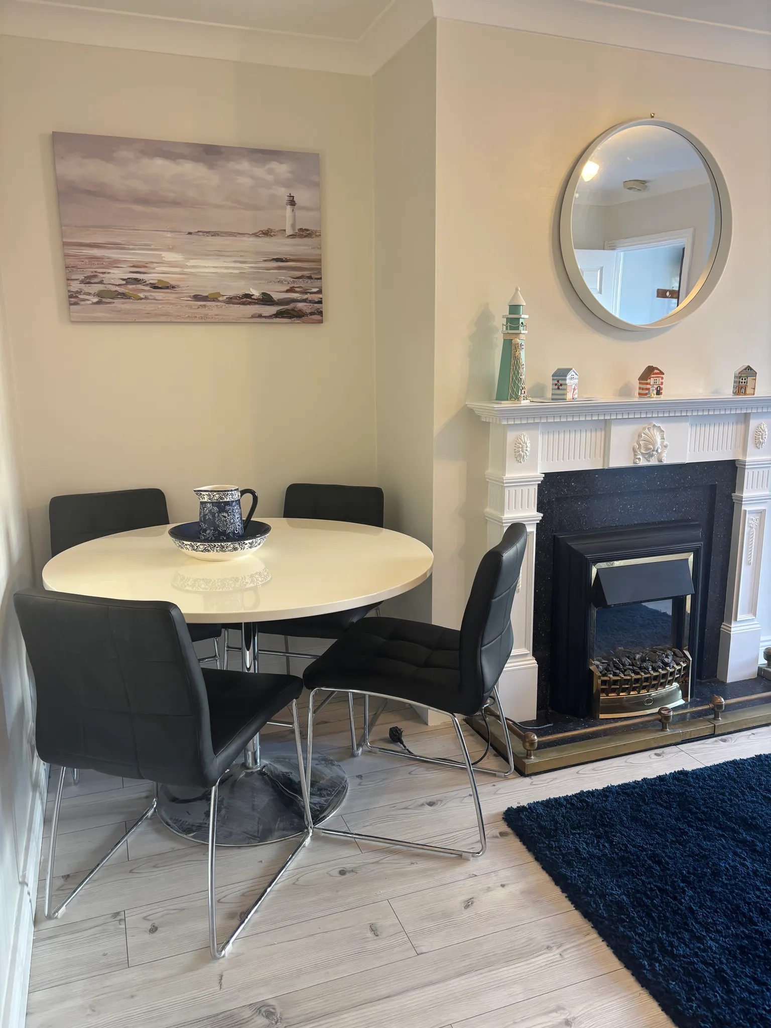 Dining room corner with a round white table, four black chairs, a blue patterned pitcher and bowl on the table, a white fireplace mantel with small decorative houses and a lighthouse, and a round wall mirror and seascape