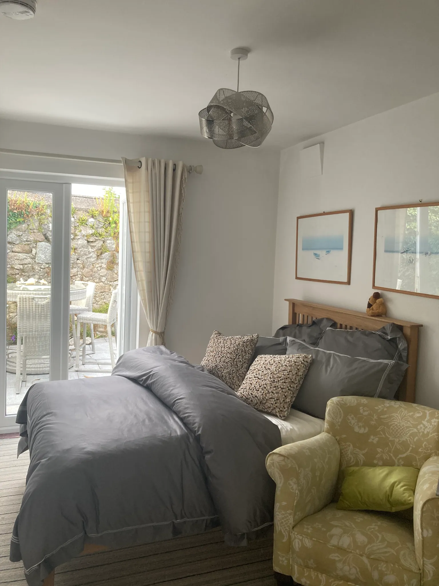 Bedroom with a double bed covered in dark bedding, patterned throw pillows, a wooden headboard, an upholstered armchair, curtains and a sliding glass door leading to an outdoor patio.