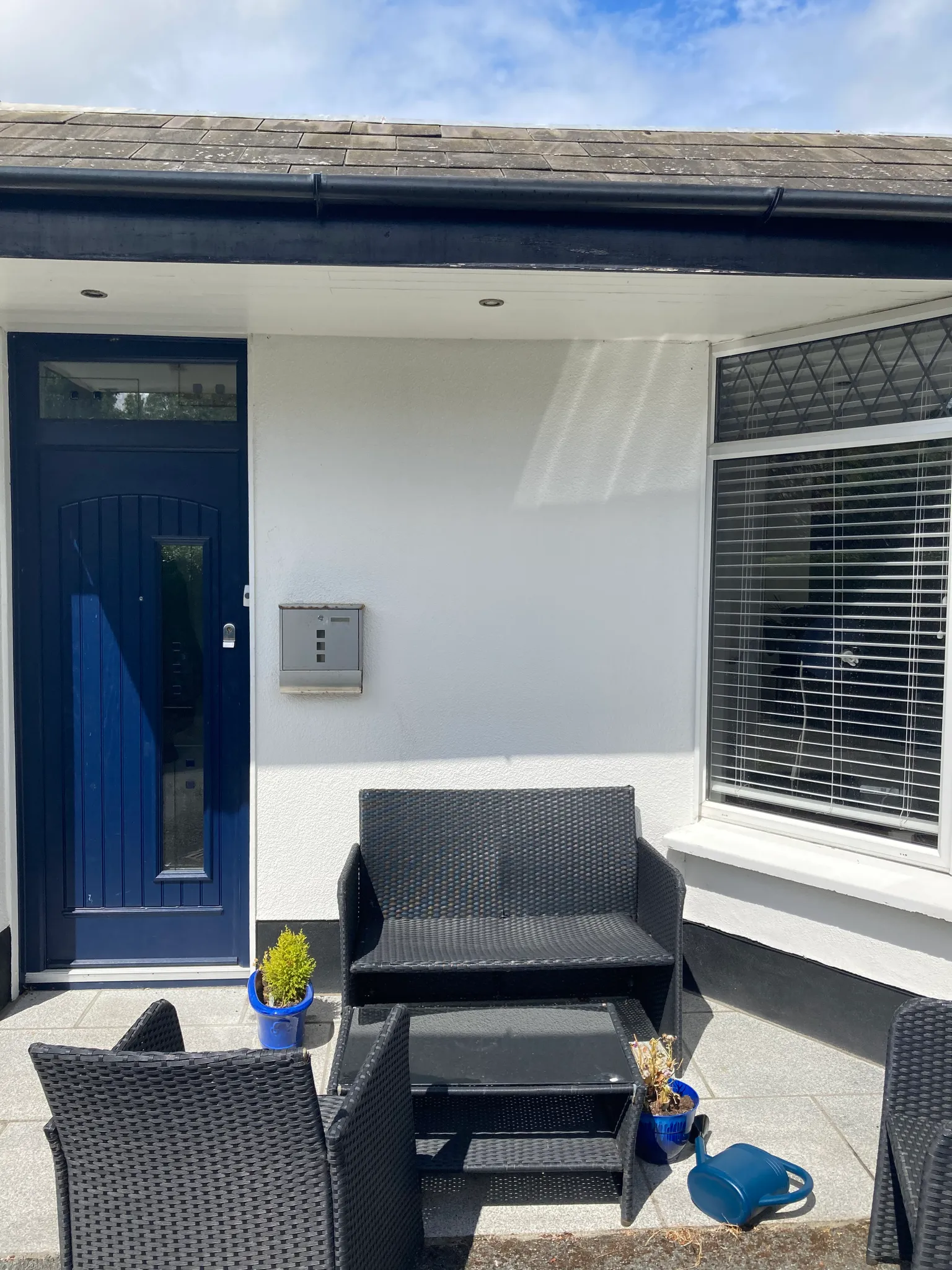 Exterior entryway with a blue front door, wall-mounted mailbox, adjacent window with blinds, and outdoor wicker seating on a small patio