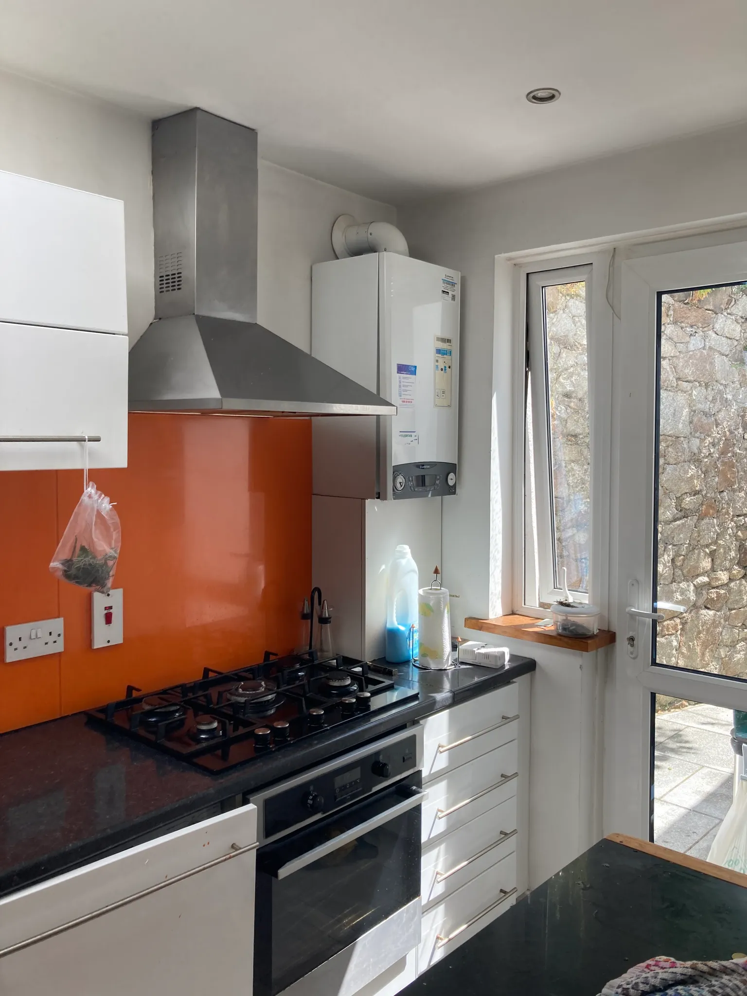 Kitchen corner with a gas hob and oven, stainless steel extractor hood, orange splashback, white cabinets, a countertop with cleaning bottles and a paper towel holder, and a windowed door to the right.
