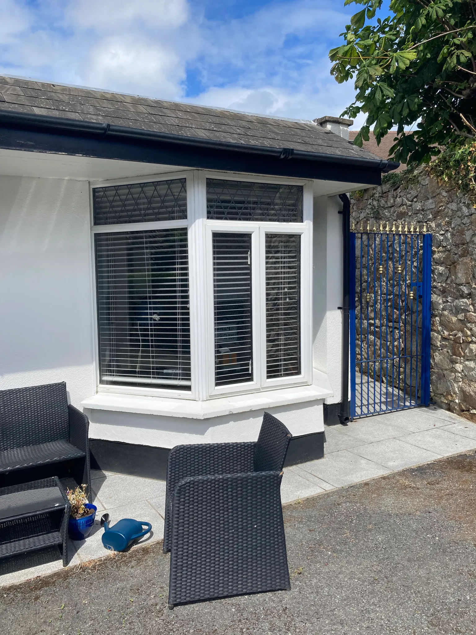 Exterior view of a bay window with closed blinds on a white wall, small paved patio with wicker chairs, blue metal gate and stone wall at right.