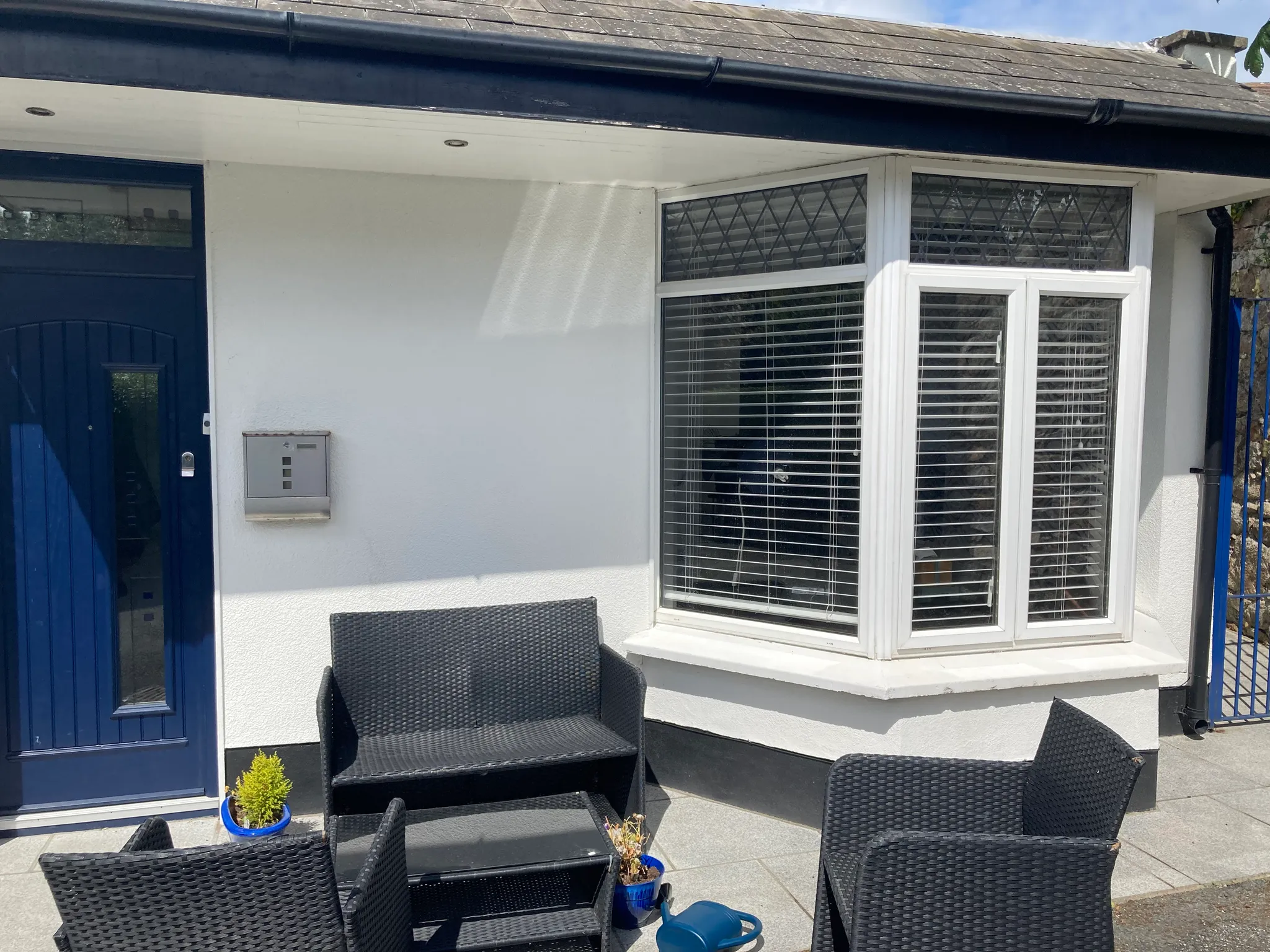 Exterior front entrance with a blue door, a white bay window with blinds, a mailbox on the wall, and dark wicker outdoor seating on a small patio.