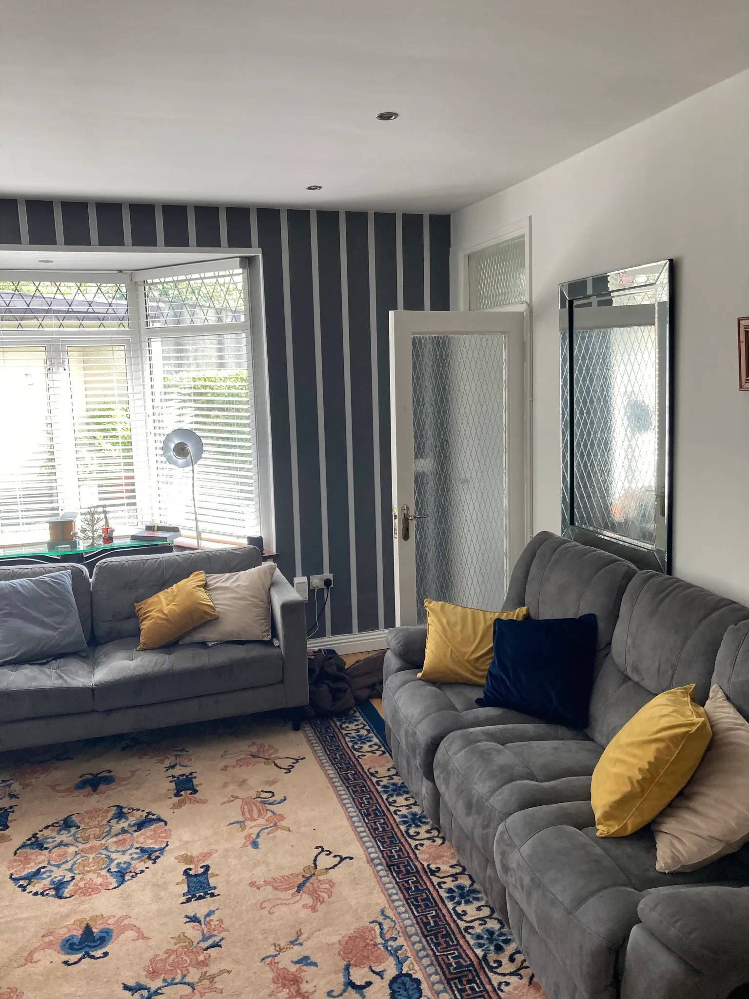 Living room with two grey sofas with yellow and blue cushions, a bay window with blinds, patterned rug, floor lamp, and a wall mirror.